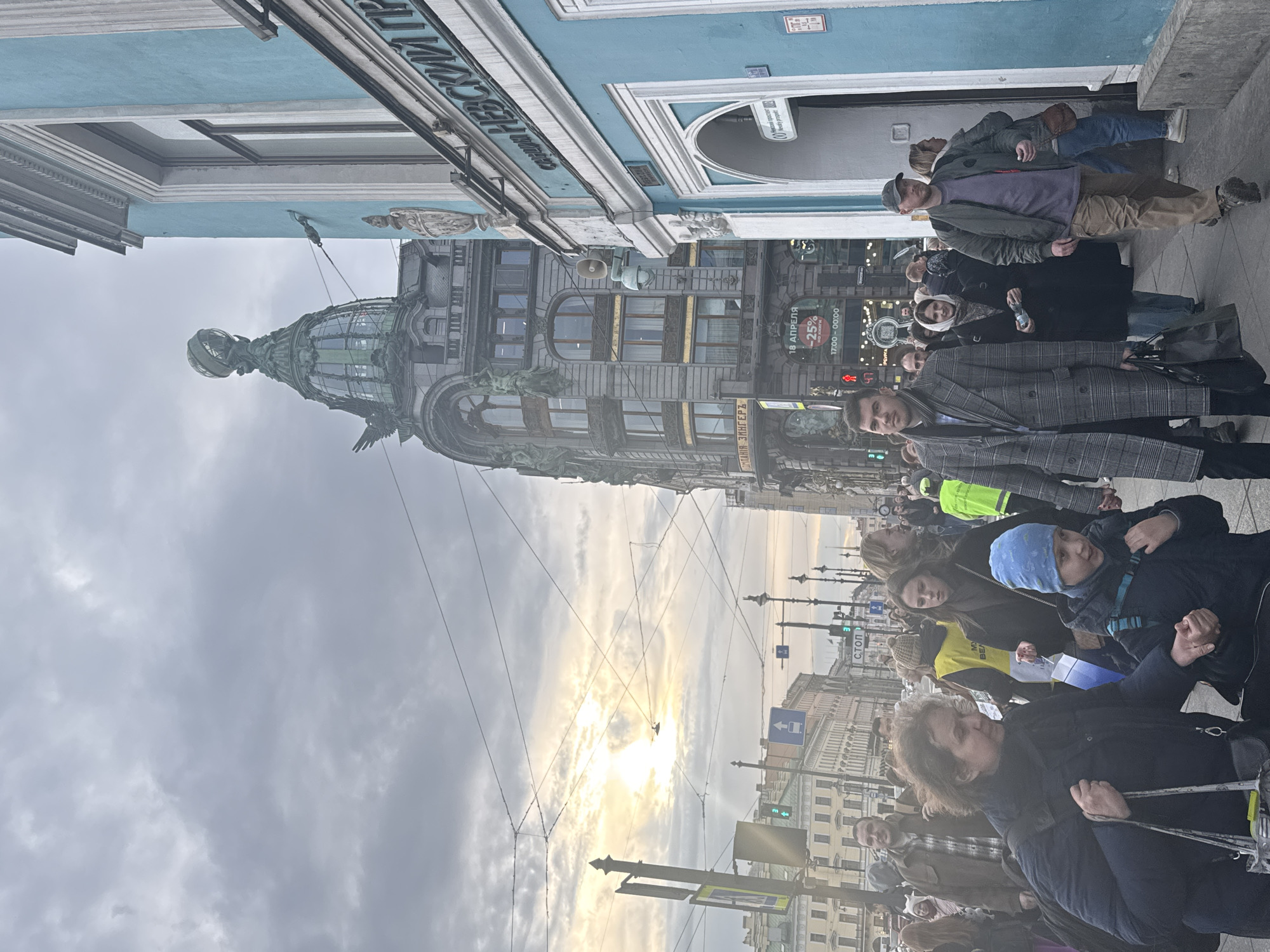 A street scene in Saint Petersburg, Russia, features the Singer House (also known as the Dom Knigi, or House of Books) with its distinctive glass dome visible against a cloudy sky. A crowd of people walks along the sidewalk in the foreground, wearing winter clothing. Overhead wires intersect the sky. Traffic signals and street signs are visible in the distance. The architecture of other buildings lines the street.