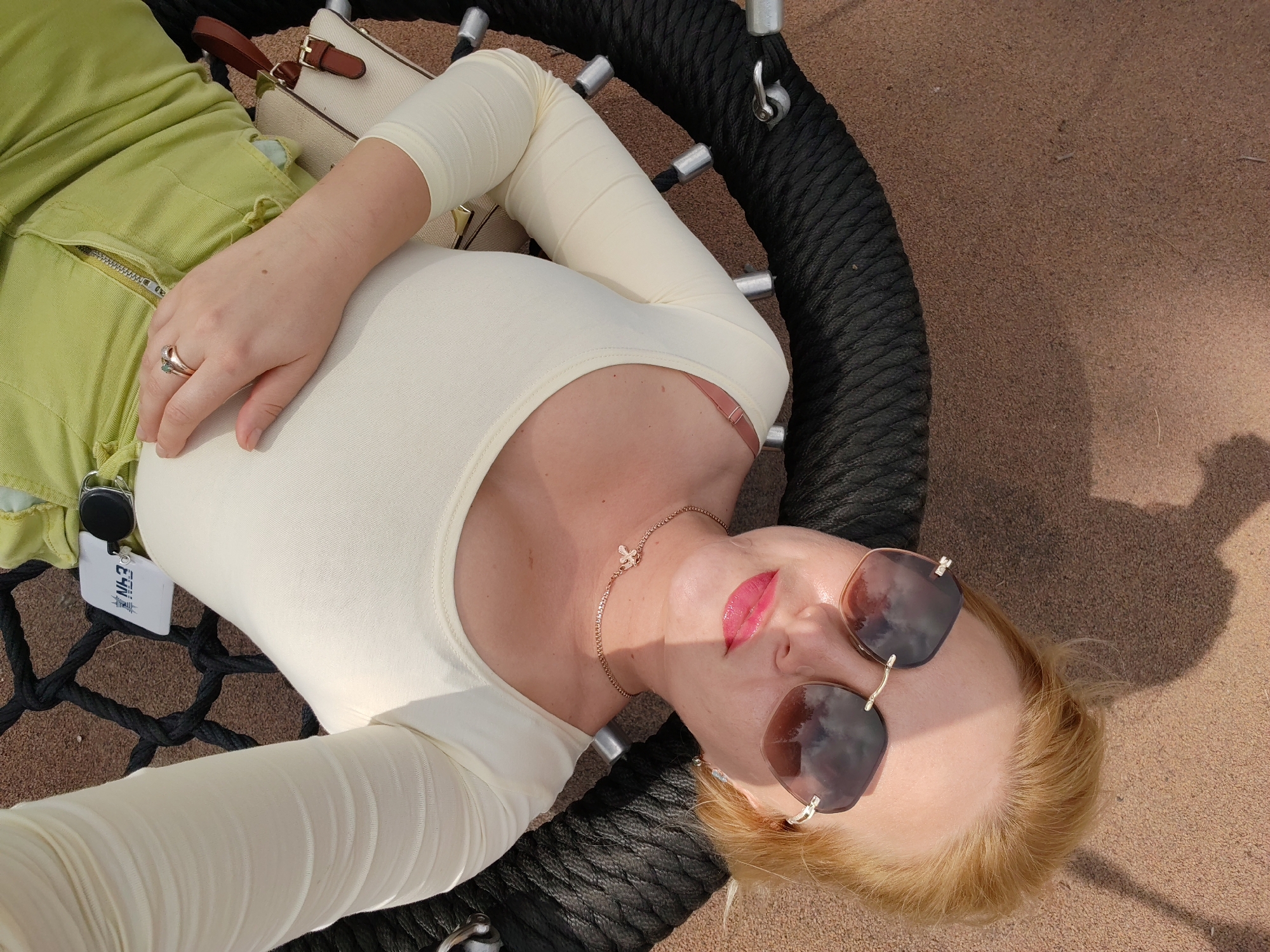 The image shows a woman lying on a swing. She is wearing a light-colored long-sleeved shirt, light green pants, and sunglasses. She has on a ring and a necklace with a bee pendant. Her lips are painted with a bright pink lipstick. A white purse is visible near her left arm. The swing appears to be made of black rope and is suspended over a sandy surface. The woman is looking up at the camera, and her face is partially illuminated by sunlight, casting a shadow on the sand. The setting appears to be outdoors, possibly in a park or recreational area. There is no specific information in the image that would allow for its location to be identified as Izhevsk, Russia.