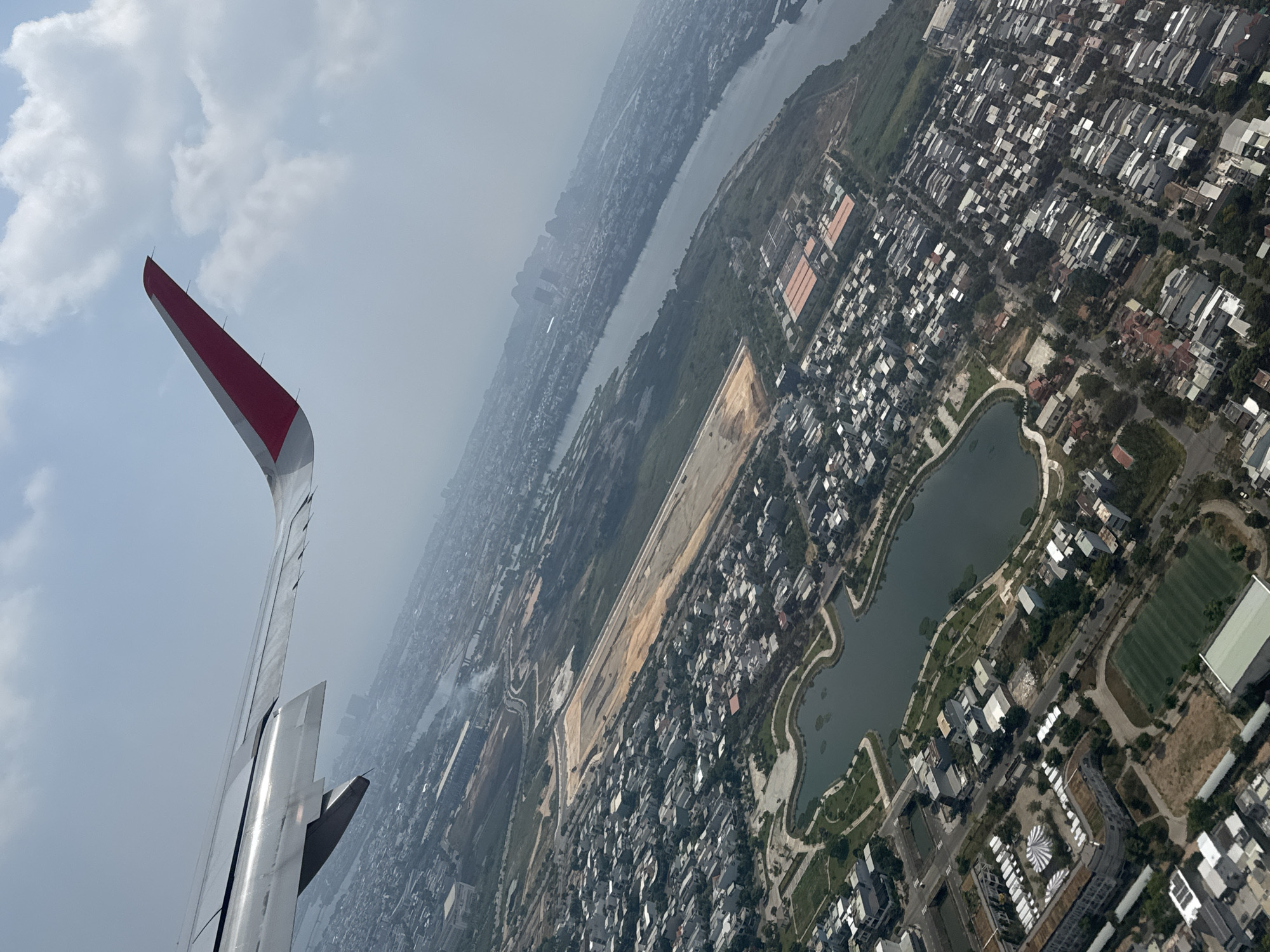 The image shows an aerial view from an airplane over a city in Đà Nẵng, Vietnam. The wing of the plane is visible on the left, obscuring part of the view. In the background, a river runs through the city. The city is densely populated with buildings and some green areas. A large construction site is visible with a large rectangular, cleared area, and near the bottom-right is a body of water. The sky is overcast with clouds.