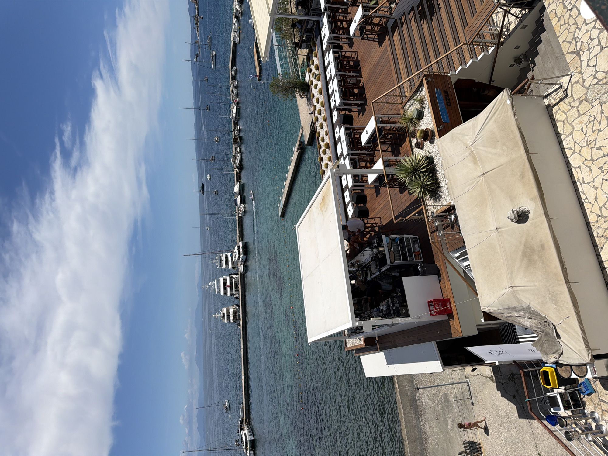 This image captures a vibrant daytime scene at a waterfront location in Corfu, Greece, showing a marina and a multi-level restaurant or bar. The perspective is from an elevated position, looking down and across the water.
In the foreground and mid-ground, a clear blue-green sea is dotted with numerous boats, including sailboats and larger yachts, either docked along piers or anchored further out. The water appears calm and inviting. Along the shore, a concrete pier or walkway extends into the water, bordered by yellow and red buoys. Mountains are visible in the hazy distance across the expanse of water.
To the right, a modern, multi-tiered restaurant or bar dominates the scene. It features extensive wooden decking, furnished with white tables and dark chairs, set under white awnings or large umbrellas, providing shade. Decorative elements include potted cacti and palm-like plants on white pebble landscaping. Metal railings delineate the different levels and stairs. A bar area is visible on one of the lower levels, with staff members tending to it.
On a concrete platform below the restaurant, one person wearing a swimsuit is visible, standing near some outdoor furniture and bins (yellow and blue). Shadows are cast across the platform, indicating bright sunlight. The sky above is a clear blue with scattered white, wispy clouds.
Visible text includes "AZUR" on a blue sign attached to the wooden structure of the restaurant, and "Coca-Cola" on a red crate. A partially obscured white banner also shows what appears to be "RS". The overall impression is one of a sunny, relaxed, and active coastal setting.
