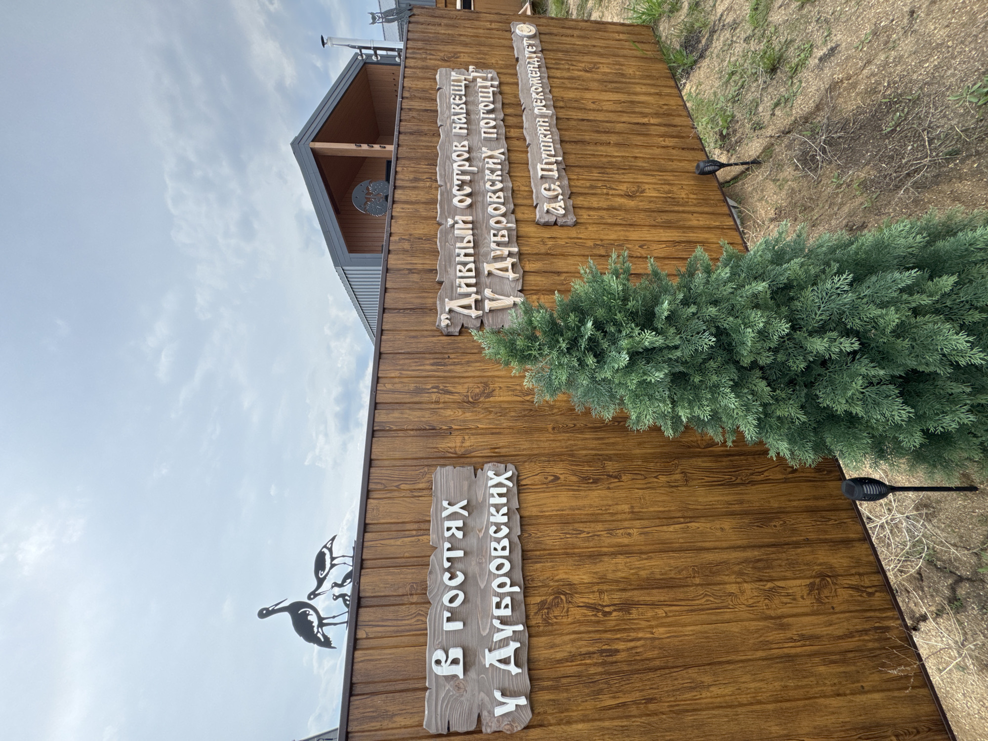 The image depicts a wooden fence with two signs and decorative metal bird silhouettes on top. The left sign reads "В ГОСТЯХ У ДУБРОВСКИХ" (Visiting the Dubrovskys). The right sign contains text in quotes: "“Дивный остров навещу У Дубровских погощу!”" (I will visit the wondrous island, I will visit the Dubrovskys!). Below this, another line reads "А.С. Пушкин рекомендует" (A.S. Pushkin recommends), with a smiling emoji. A small evergreen tree is planted in front of the fence. In the background, a modern building with a gabled roof is visible. The scene appears to be outdoors, likely in a rural or semi-rural setting. The location is identified as Khuzhir, Russia.