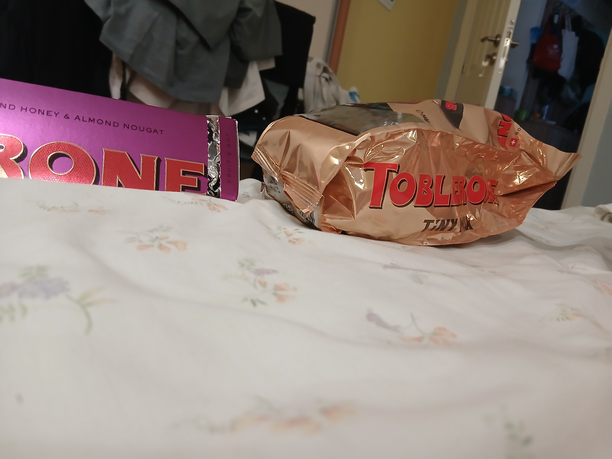The image depicts a close-up view of two chocolate bars, one Toblerone and one Cadbury, resting on a white, floral-patterned surface. The Toblerone bar, partially opened and crinkled, is in the foreground, with its distinctive red lettering visible. The Cadbury bar, also with visible red lettering, is positioned to the left, with its purple packaging and the words "ND HONEY & ALMOND NOUGAT" legible. The background is blurred, showing a doorway with a handle and a glimpse of clothing hanging on a rack. The scene appears to be set indoors, possibly on a bed or a soft surface, with soft lighting. No specific location in Pleven, Bulgaria can be determined from the image.