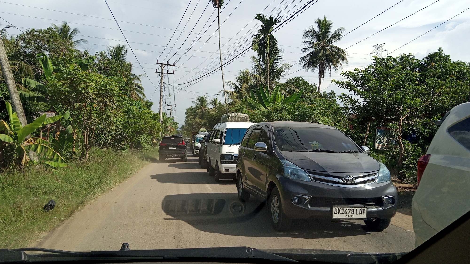 The image depicts a traffic jam on a rural road lined with lush green trees and vegetation, characteristic of the outskirts of Blang Mee, Indonesia. The road is unpaved and dusty, with a clear sky overhead dotted with clouds. Several vehicles are visible, including a dark gray Toyota Avanza in the foreground, a white van, and other cars further down the road. The Toyota Avanza has a license plate that reads "BK1478 LAP" with "03-31" below it. The vehicles appear to be stationary, indicating a traffic standstill. Utility poles with overhead wires run along the side of the road, and palm trees are interspersed among other foliage. The scene suggests a common occurrence of traffic congestion in a developing area with natural surroundings.