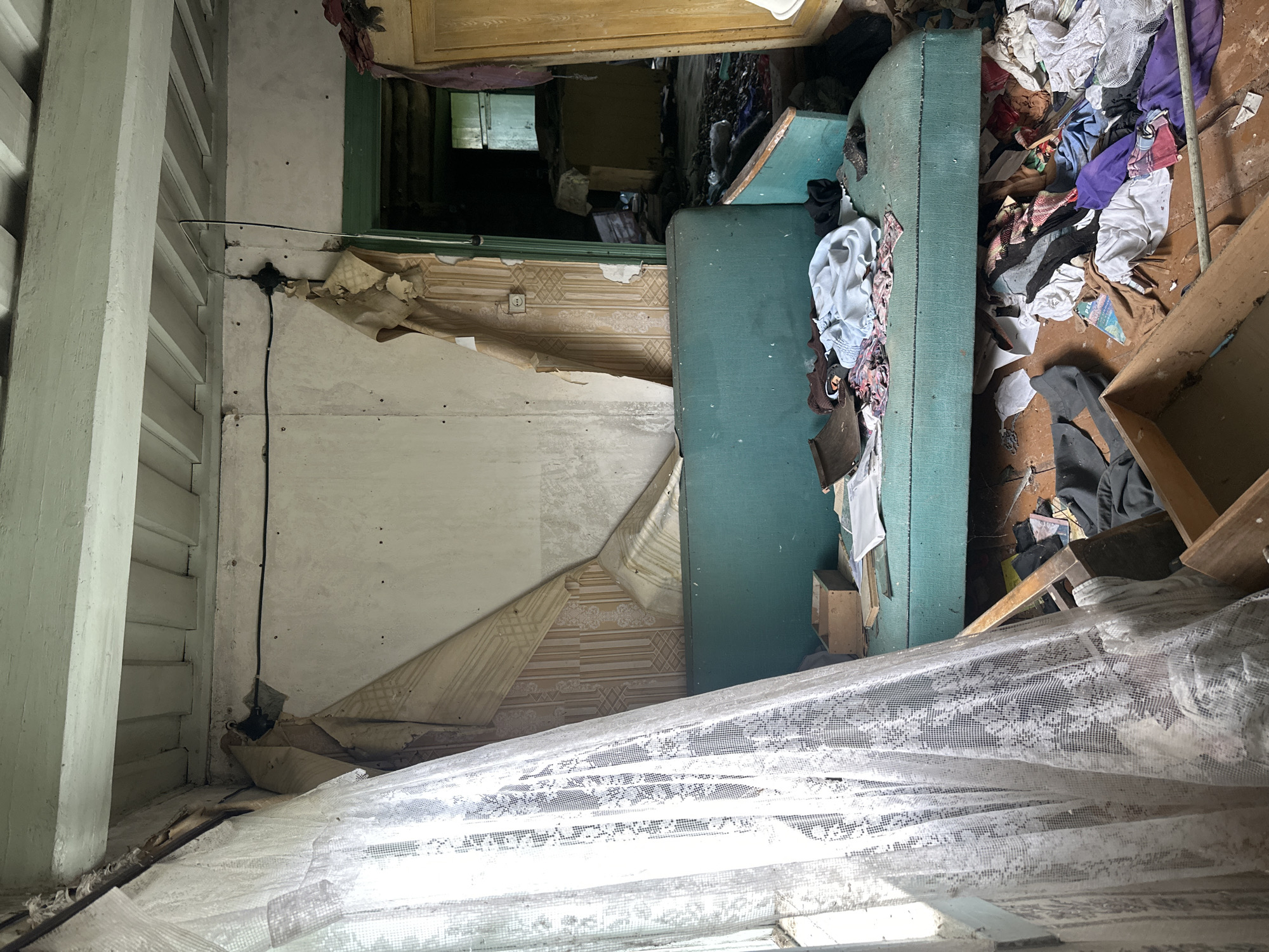 The image depicts the interior of a room in a state of disarray and neglect, likely an abandoned or neglected dwelling. The scene is dominated by clutter, including a large teal-colored sofa or mattress covered with various items of clothing and fabric. The walls are partially covered in peeling wallpaper, revealing older layers beneath, and in some areas, the plaster is exposed and damaged.
Several objects are scattered across the floor and on surfaces, contributing to the sense of chaos. These include more clothing, wooden planks, and what appear to be pieces of furniture or structural elements. A white lace curtain hangs in the foreground, partially obscuring the view and suggesting a window is present. Electrical wires are visible, with one black cable running down a wall and connected to a junction box.
In the background, a doorway leads to another space, where more debris and darkness are apparent. A sliver of light can be seen coming from what might be a distant window or opening. The overall impression is one of decay and abandonment. There is no discernible text in the image. The lighting suggests it is daytime, but the condition of the room evokes a somber or melancholic atmosphere. The location context of Відзаўскі сельскі Савет, Belarus, points to a rural setting within the country.