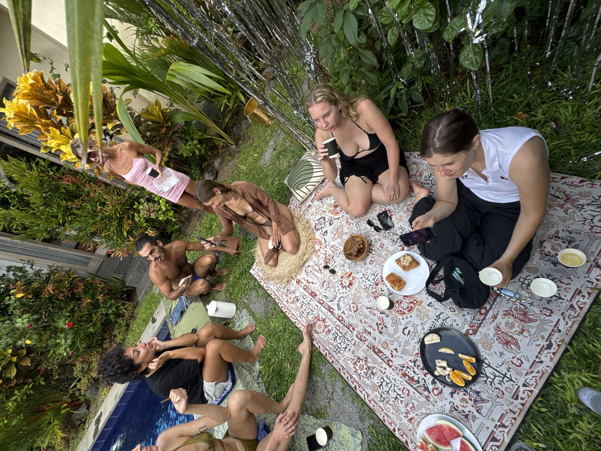 In North Kuta, Indonesia, a group of people are gathered outdoors, possibly in a backyard setting, seemingly for a casual get-together or picnic. The camera's perspective is angled.
People:
- A woman in a pink dress holding a container.
- A woman in a brown outfit sitting cross-legged.
- A shirtless man is squatting, holding a phone.
- A woman wearing a black bathing suit.
- A woman in a white shirt and black pants is seated on the ground.
- Several other individuals are near a pool.
Objects:
- A patterned blanket spread out on the grass.
- A variety of food items, including pastries, fruit (watermelon, bananas, oranges).
- Cups and bowls scattered on the blanket.
- Mobile phones in use.
- A pool with visible water.
- Plants and trees, suggesting a tropical environment.
- Decorative silver strips in the background.
Actions and Interactions:
- People sitting, eating, drinking.
- Some are looking at their phones.
- Conversation among those in close proximity.
Environment:
- A lush green lawn serves as the central area.
- Dense vegetation.
- The presence of a pool suggests a residential or resort setting.
- The tropical climate is implied by the surrounding plants.