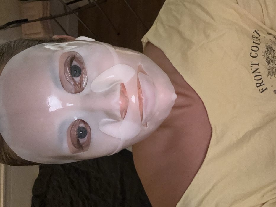 A person wearing a sheet mask is visible. The mask is white and covers most of the face, with cutouts for the eyes, nose, and mouth. The person has fair skin and blue eyes. They are wearing a yellow t-shirt with the words "FRONT COURT" printed on it. The image is slightly rotated. A dark, possibly furry object is visible in the lower left corner. A metal rack is visible in the background.