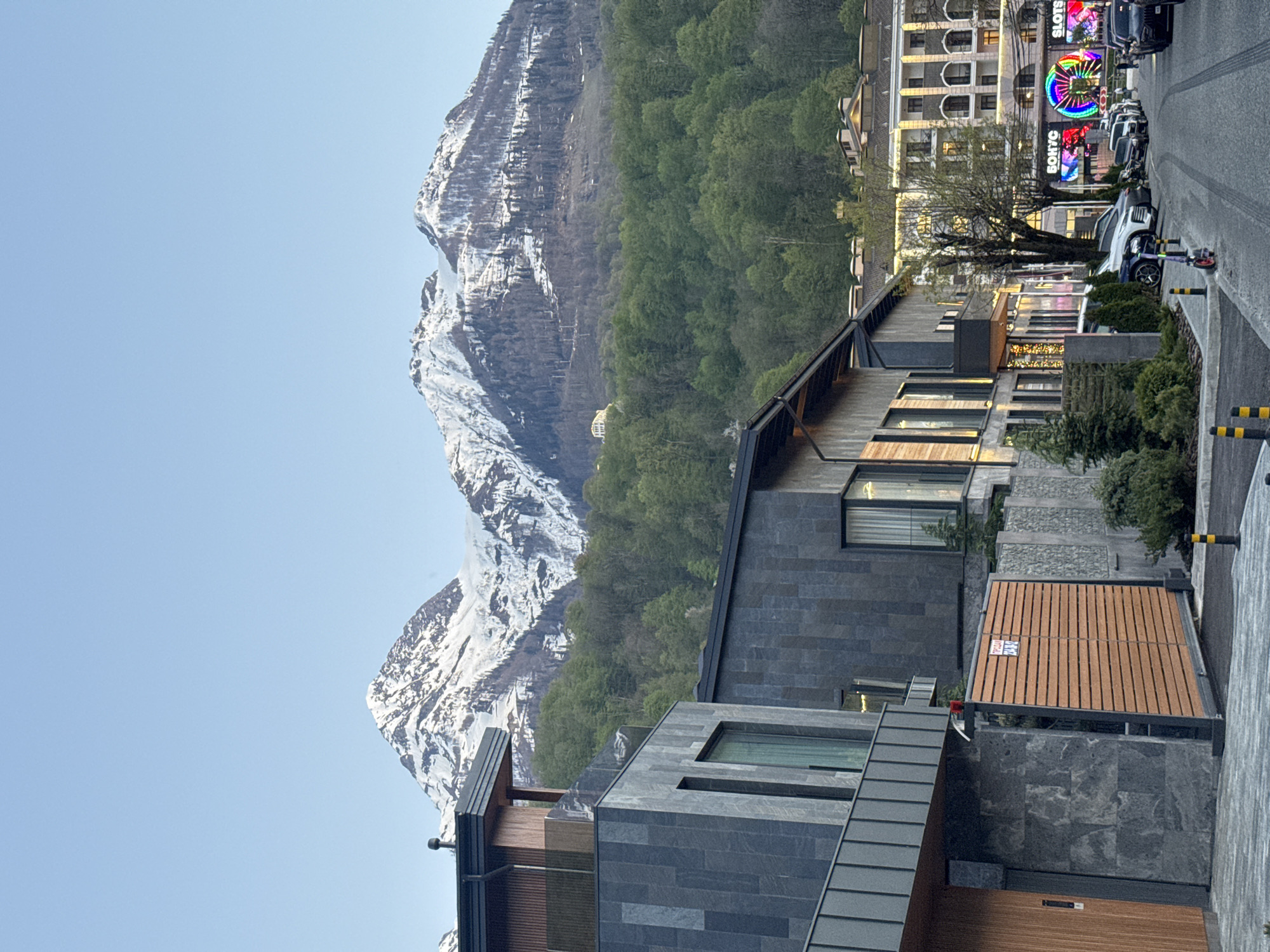 The image shows a view in Estosadok, Russia, featuring a blend of modern architecture and natural scenery. Snow-capped mountains dominate the backdrop. A modern building with a combination of dark gray stone and light-colored wooden siding occupies the foreground. The exterior includes multiple windows and sleek architectural lines. Adjacent to the building is a paved area and a road. On the road, there are several parked vehicles. Further in the distance, commercial buildings are visible, including one with a brightly lit "SLOTS" sign. The sky is clear blue.