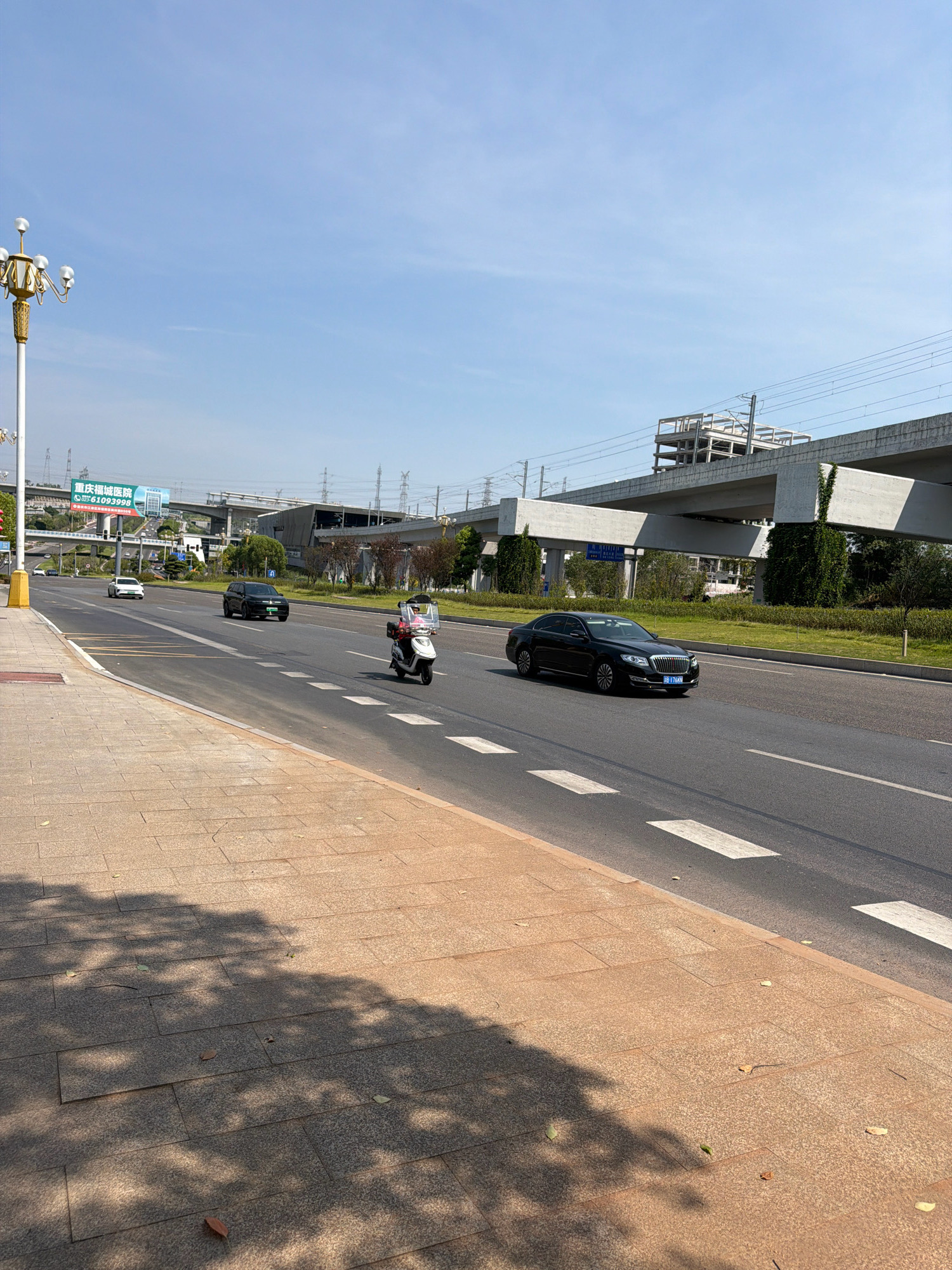 In Jiangjin, China, this outdoor urban scene captures a bright, sunny day with a clear blue sky. The perspective is from a sidewalk made of textured reddish-brown and grey rectangular tiles, casting dappled shadows from overhead trees onto the ground. The sidewalk borders a multi-lane asphalt road with visible white dashed lines and pedestrian crossing markings.
On the road, a white electric scooter, ridden by a person wearing a light-colored shirt and a pink mask, is traveling away from the viewer. To the left of the scooter, a dark-colored SUV is also moving in the same direction. Further down the road, a white sedan is visible. On the right side of the road, a black luxury sedan is driving towards the viewer.
In the background, an elevated concrete overpass with train tracks runs across the scene, supported by large pillars. Behind the overpass, several large power line structures and pylons are scattered across the landscape. A billboard with Chinese text and a phone number is positioned to the left of the overpass. A traffic light pole stands near the billboard. The overall environment suggests a modern urban area with significant infrastructure development, including transportation and power lines.