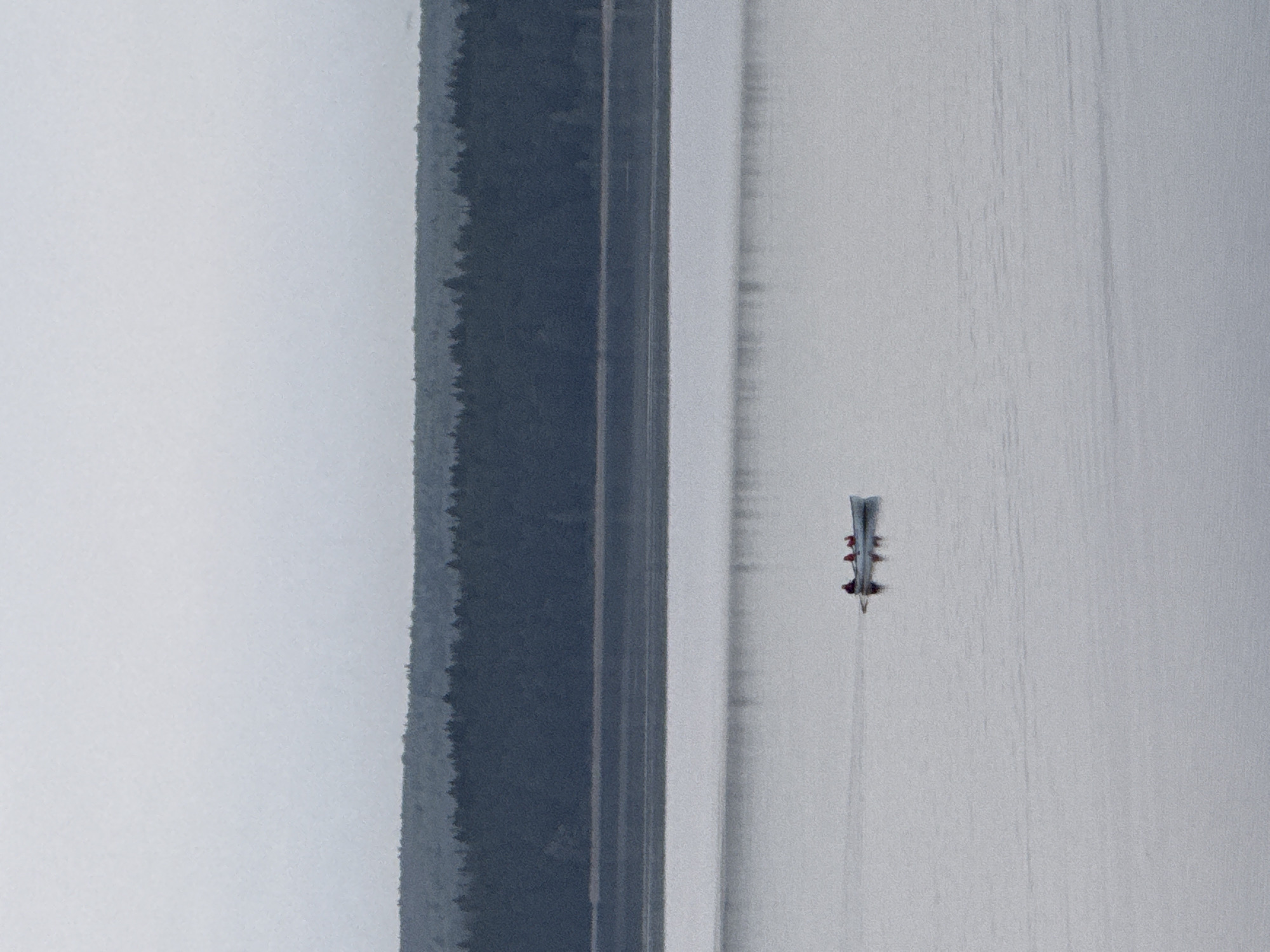 An aerial view shows a rowing shell on a body of water next to a road and forested area. The shell is moving away from the viewer, leaving a wake. People are visible in the shell. The scene is in Votkinsk, Russia.