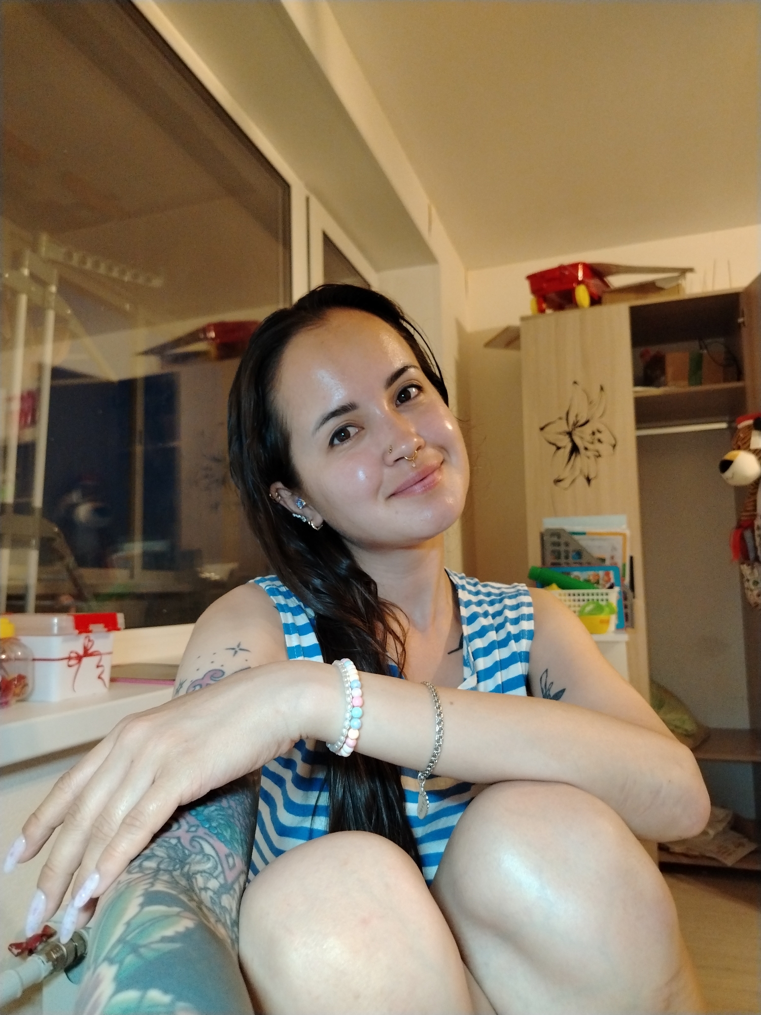 A woman with tattoos sits indoors. She has long dark hair, a nose ring, and multiple earrings. She is wearing a blue and white striped top and two bracelets on her right wrist. Her left arm, heavily tattooed with a blue and green design, rests on her knees, which are drawn up towards her chest. Her fingernails are long and painted white.
Behind her, a window reflects an outdoor scene. To the right is an open wardrobe with items on the shelves. On top of the wardrobe is a red toy car. The location is likely Noviy, Russia based on the original prompt.
