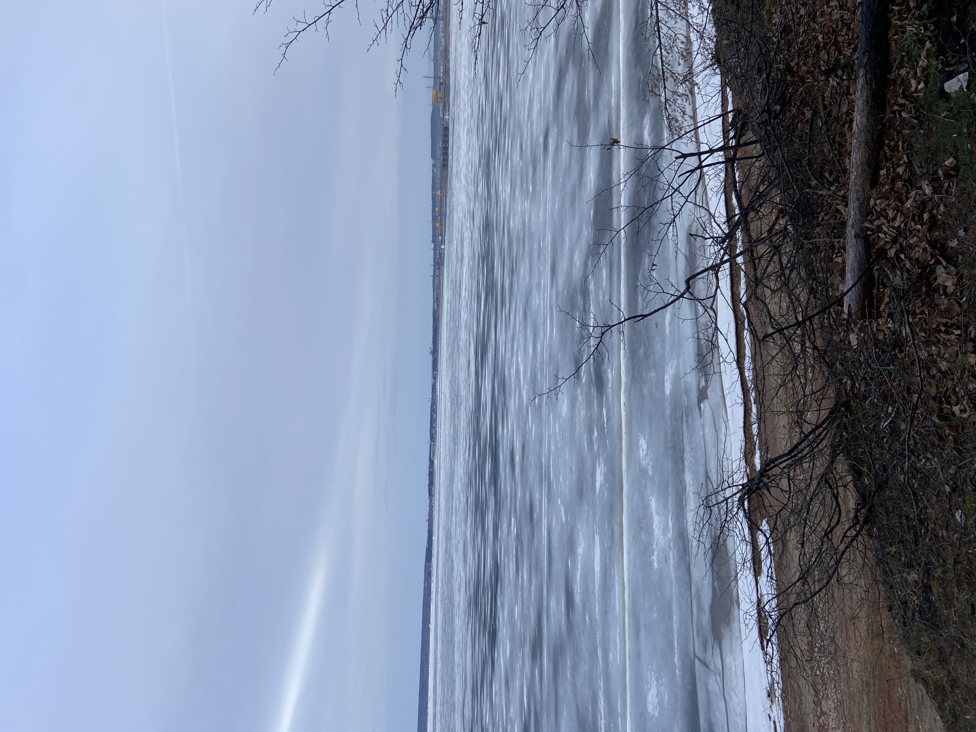 The image depicts a frozen body of water, likely a river or lake, adjacent to a muddy or sandy embankment covered with dry leaves and bare tree branches. In the distance, a cityscape is visible, identified as Noviy Urengoy, Russia. The sky is overcast and pale blue. The ice on the water appears textured and patterned, suggesting varying degrees of freezing or thawing. A few bare tree branches are visible near the edge of the frame. No people are visible.