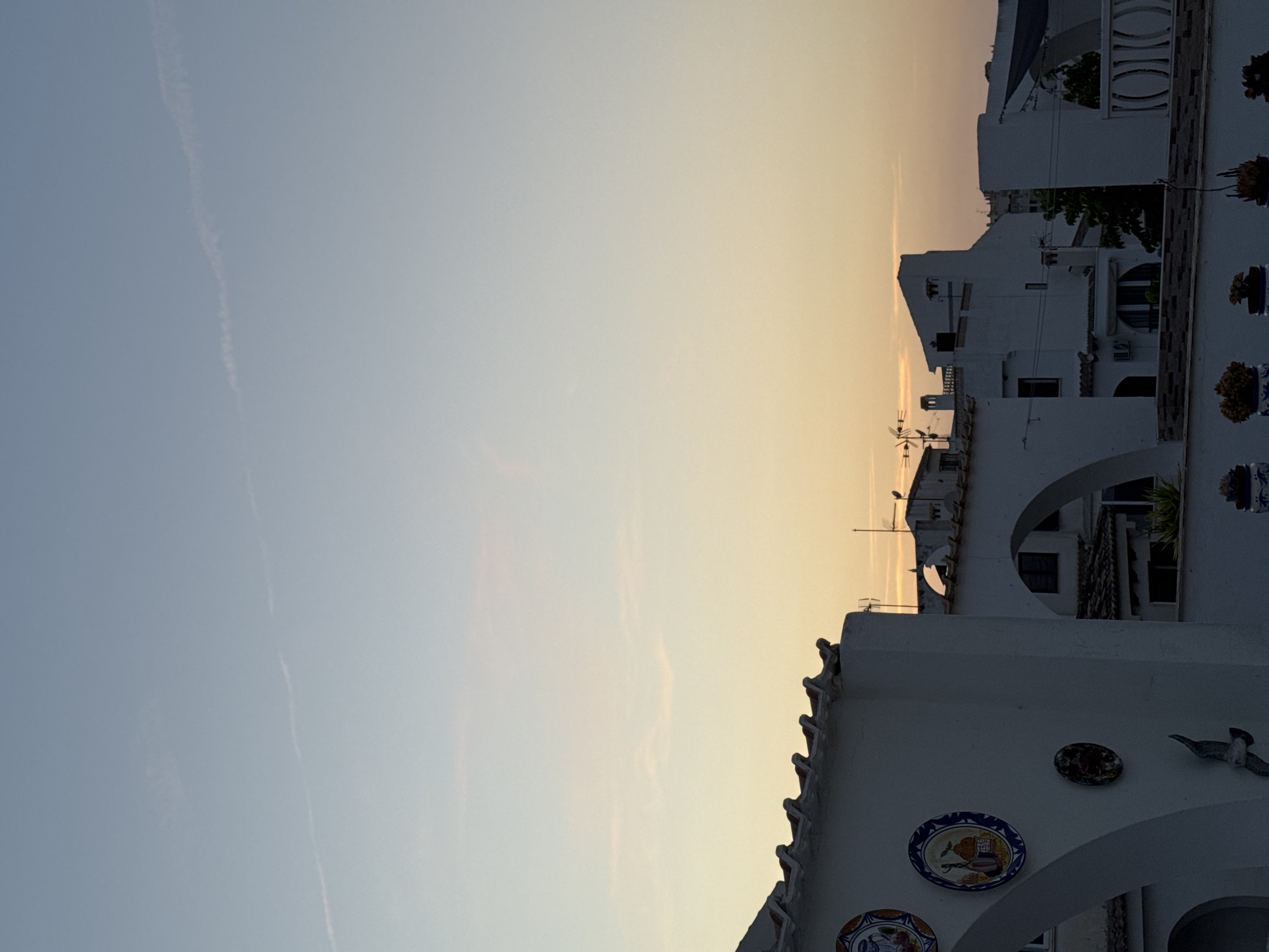 The image shows a view of white buildings in Torrevieja, Spain, under a pale sky with a faint sunset glow. The foreground features a white building with decorative ceramic plates on its facade and a bird-shaped ornament. Arched doorways are visible on this building. Behind it, other white buildings with tiled roofs and some with visible antennas are seen. The sky transitions from a light blue at the top to a soft yellow and orange near the horizon where the sun is setting. There are no people or animals clearly visible in the scene. The architecture suggests a Mediterranean style, common in coastal towns like Torrevieja.