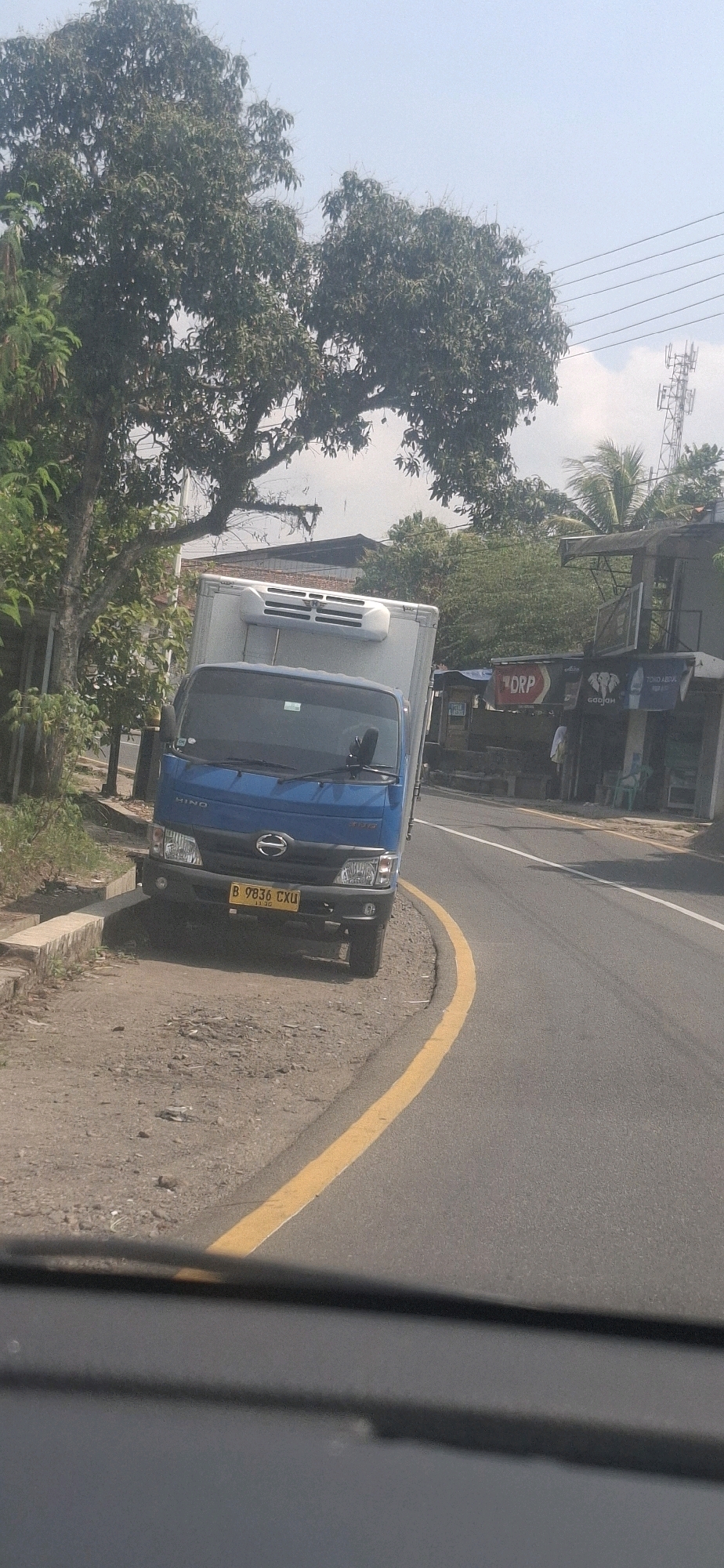 In Susukan, Indonesia, a blue HINO truck with a white refrigeration unit is parked on the shoulder of a paved road. The truck has a yellow license plate with the inscription "B 9836 CXU". The road is bordered by a yellow line on the right and a dirt shoulder with a concrete curb on the left. Lush green trees line the left side of the road, and buildings with visible signage, including what appears to be "ORP" and a depiction of elephants, are present on the right side. A cell tower is visible in the distance on the right. The sky is clear and blue, suggesting a daytime scene.