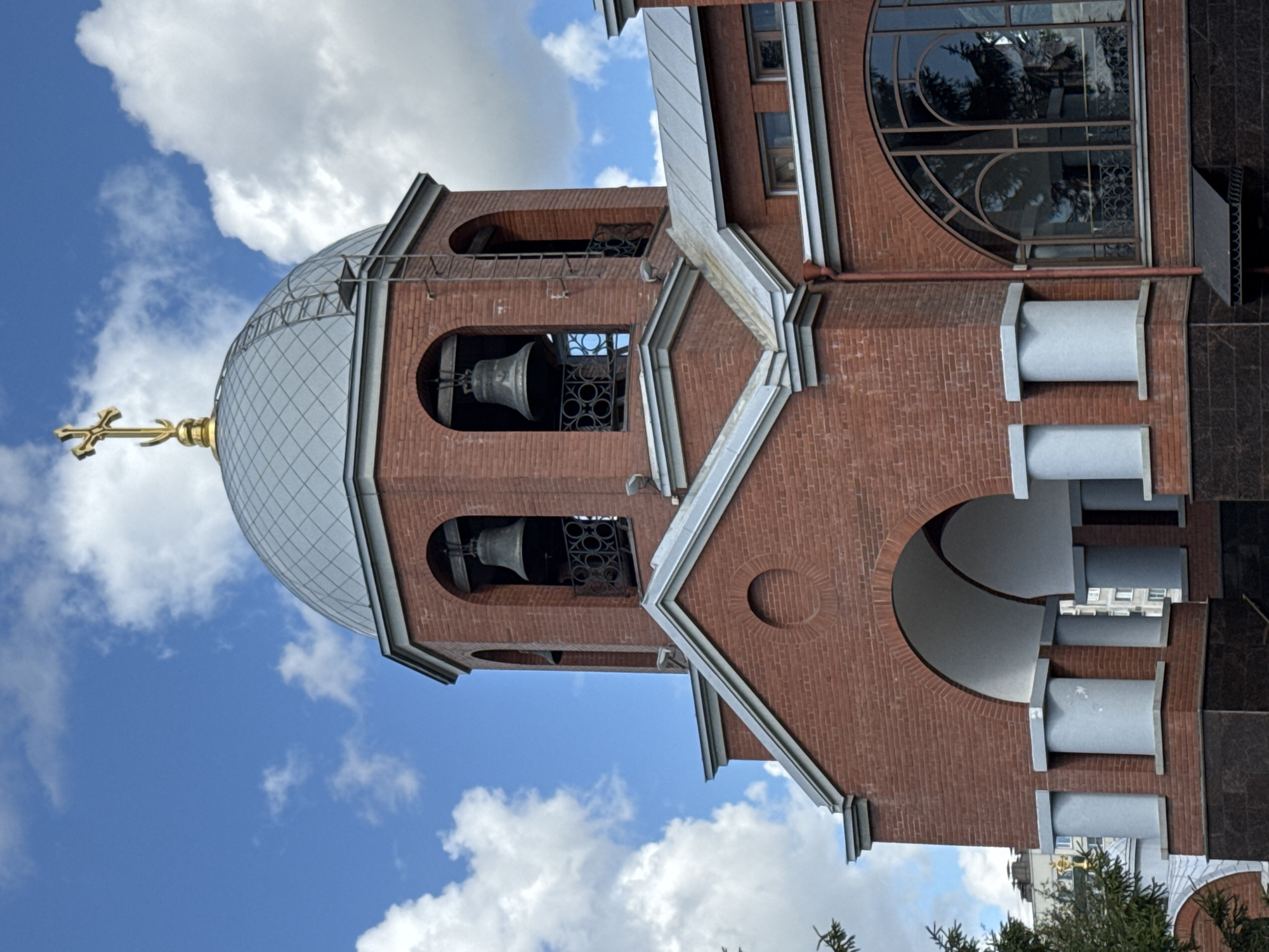 The image shows a close-up, eye-level, exterior view of the bell tower and main facade of the Church of the Resurrection of Christ, located at the Moscow Triumphal Gate in Saint Petersburg, Russia. The church is constructed primarily of red brick.
The focal point is the bell tower, which is topped with a silver-colored dome and a golden Orthodox cross. Arched openings in the tower reveal two large bells suspended within, each with black metal decorative grating below. A metal ladder extends up the side of the tower, reaching towards the dome.
Below the bell tower, the church's facade features a large, arched entrance supported by four fluted columns with grey capitals and bases. The roofline above the entrance is triangular and also has a grey metal finish. To the right of the entrance, a large, arched window with intricate metalwork is visible, reflecting trees and the sky.
The background consists of a bright blue sky with scattered white, puffy clouds. A small portion of a green evergreen tree is visible on the far left, and other buildings can be seen in the distance behind the church. The image is sharply focused, with good lighting that highlights the architectural details and textures of the brickwork and metal elements. There are no people visible in the frame.
