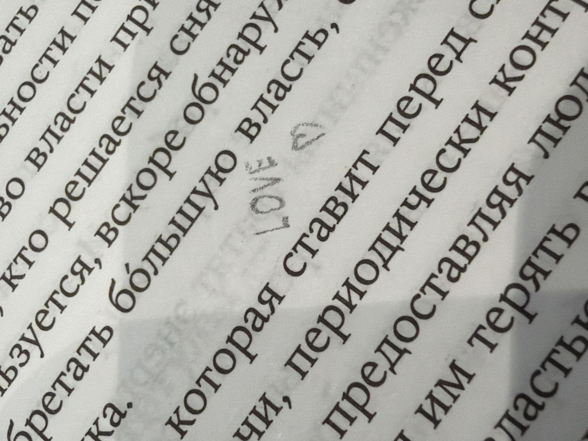 The image is a close-up shot of a page from a book written in Russian. The text is printed in a serif font and is arranged in neat rows. Overlapping one of the sentences are the words "LOVE" and a small symbol, possibly a heart, drawn in pencil. The page appears to be slightly textured. The angle of the photo is not directly perpendicular to the page, as the lines of text slant diagonally.