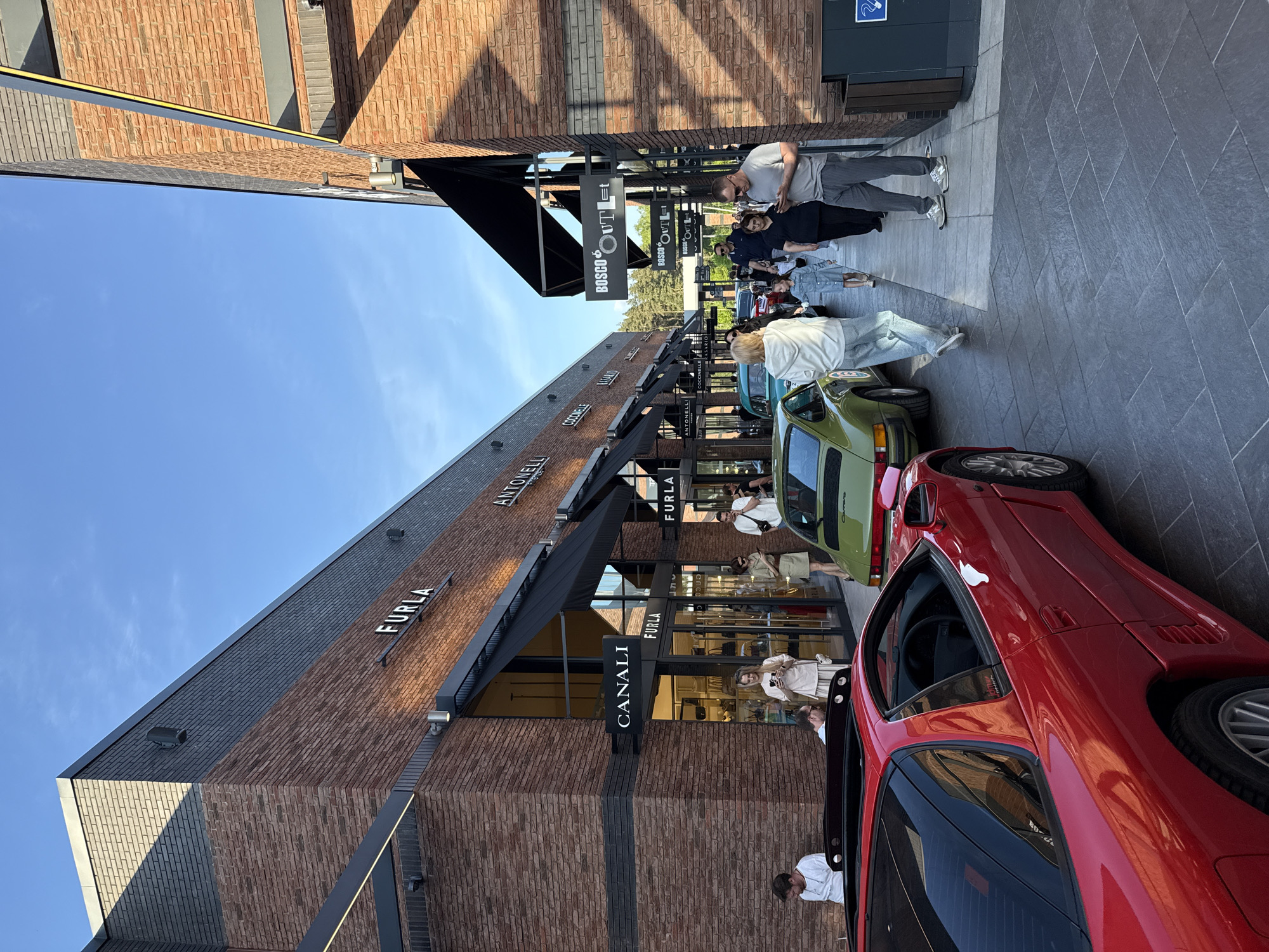 The image captures an outdoor scene in the city of Unknown, Unknown, featuring a row of retail stores. The storefronts, constructed with brick and a dark gray roof, are marked with awnings and signage for brands such as "FURIA," "ANTONELLI," "CANALI," and "BOSCO OutLEt."
Several people are walking or standing on the paved walkway in front of the stores, including those browsing shop windows. In the foreground, two sports cars, one red and one light green, are parked beside the shops. A clear blue sky appears above the buildings.