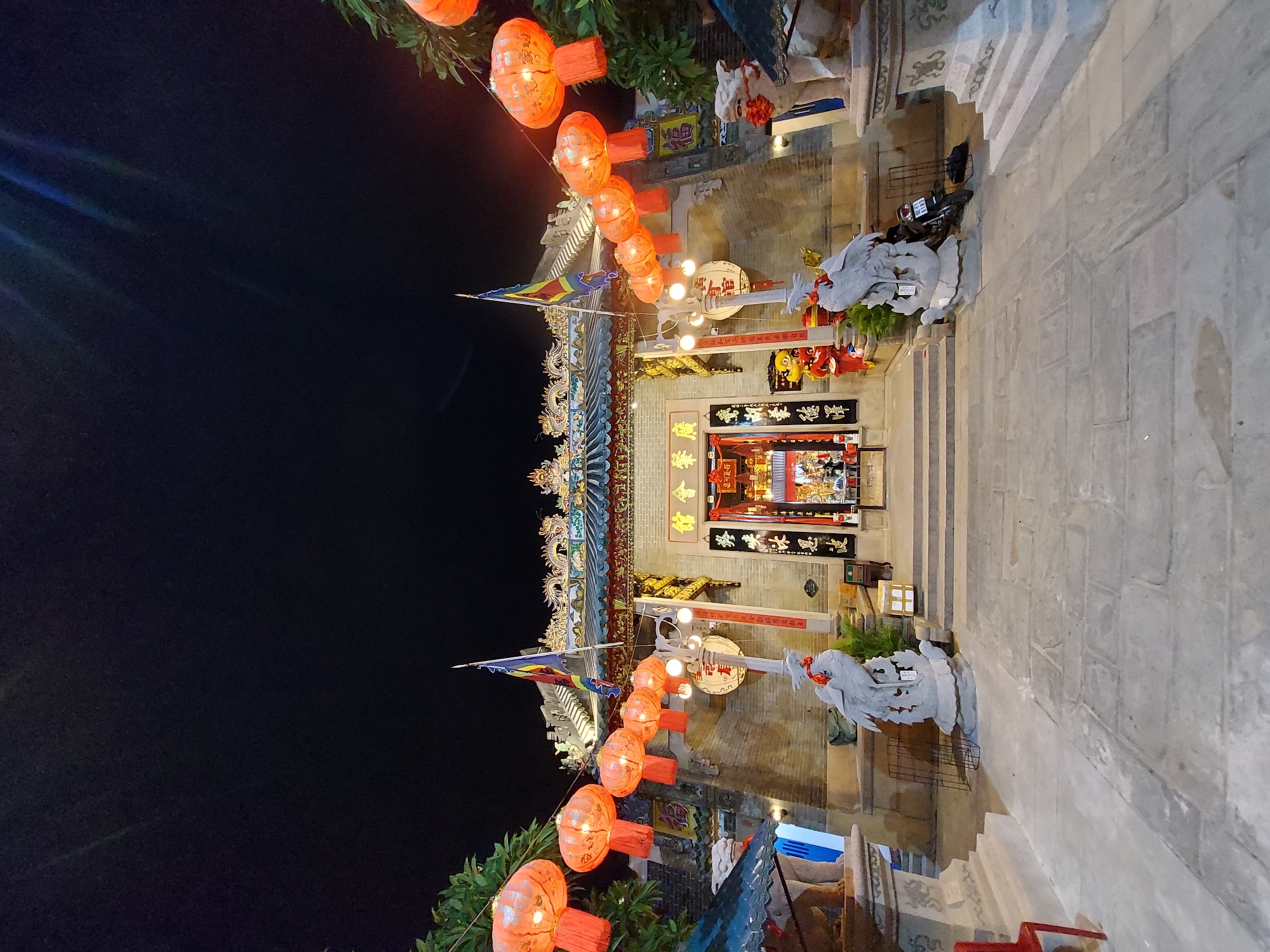 The image depicts the entrance of a Chinese temple in Hoi An, Vietnam at night. The temple is adorned with numerous red lanterns, flags, and ornate roof decorations featuring dragons. Two stone Foo dog statues flank the entrance, and a stone statue of a bird on a turtle stands in the foreground. The temple entrance is lit, revealing calligraphy on scrolls and a shrine inside. A motorcycle is parked to the right of the entrance. The ground in front of the temple is paved with large stone tiles. The sky is dark, indicating nighttime.