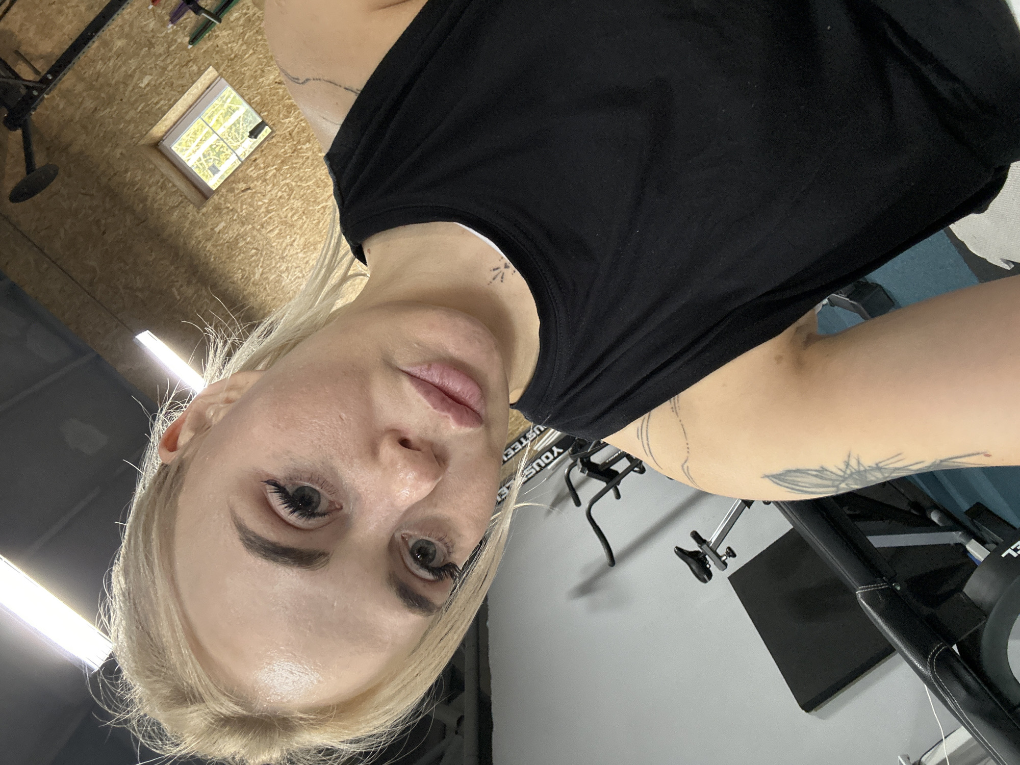 A fair-skinned person with blond hair wearing a black tank top is in an indoor space with an exercise bike. The background shows wood paneled walls with a window. Based on metadata, the location is Megion, Russia.