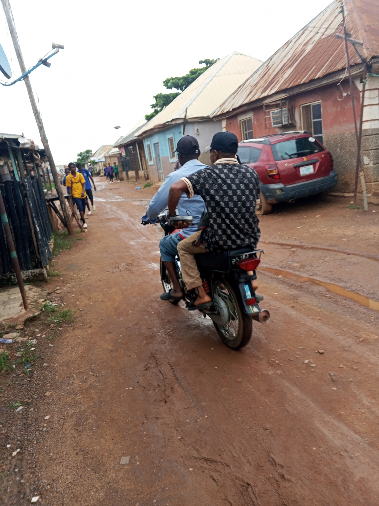 The image depicts a street scene in Abuja, Nigeria. Two men are riding on a motorcycle, with the driver in front and a passenger behind him. The motorcycle is traveling on a dirt road that appears to be wet, suggesting it may have recently rained. Buildings line the street on both sides, some with corrugated iron roofs. A red car is parked on the right side of the road. In the background, other people can be seen walking along the street. The overall impression is of a typical urban environment in a developing city.