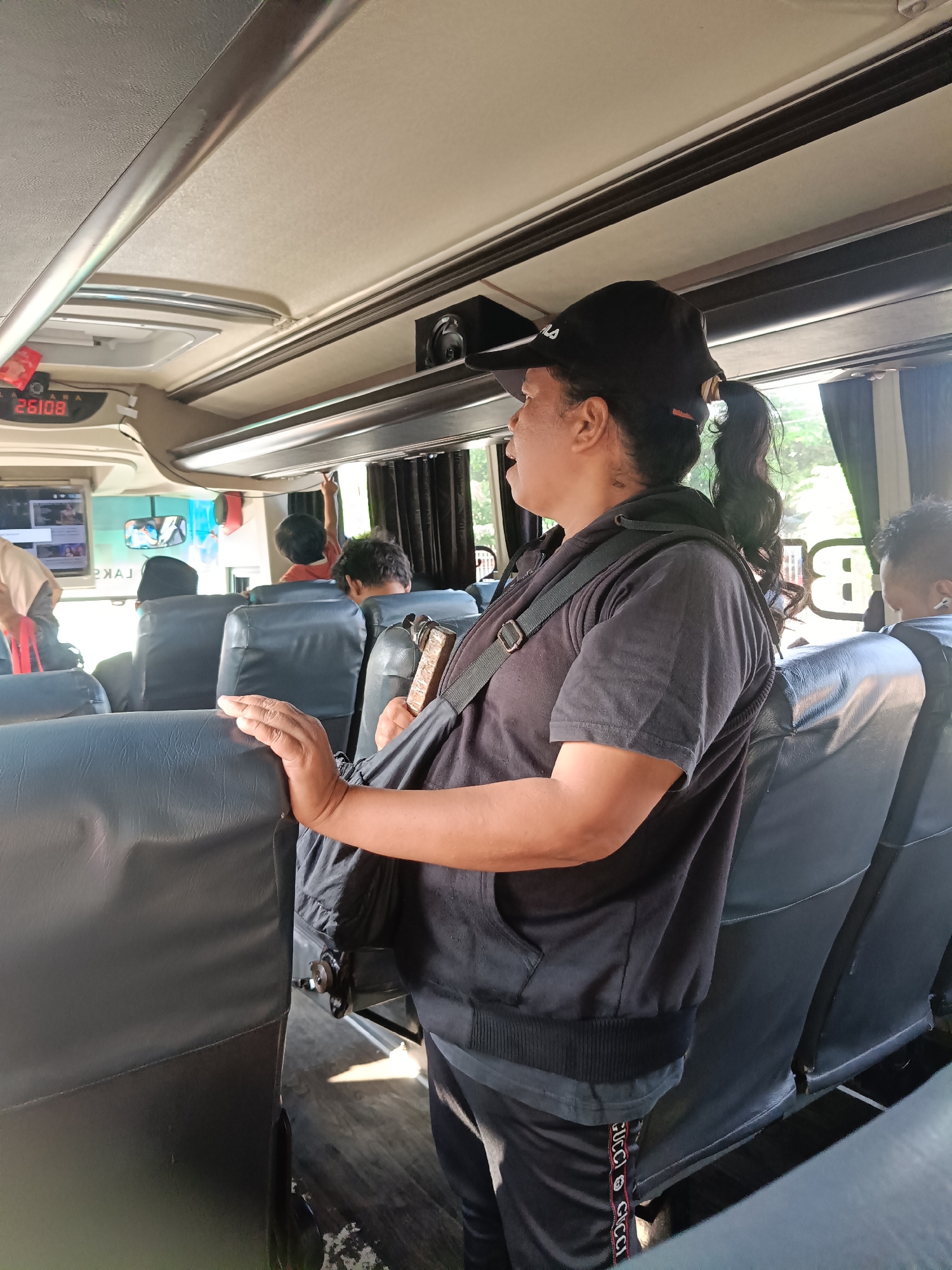A medium shot captures a woman standing inside a bus, facing right and looking out the window. She wears a black baseball cap, a black hooded jacket over a black t-shirt, and black track pants with a red and white striped logo. A dark strap crosses her chest, supporting a small, light-colored bag. Her right hand rests on the back of a dark blue seat. The bus interior features multiple rows of dark blue seats and a light-colored ceiling with overhead compartments. A digital display with the number "26108" is visible on the left. Other passengers, including a child in an orange shirt, are seated in the background. Sunlight streams in from the windows, illuminating the scene. The location is identified as Kedungrejo, Indonesia.