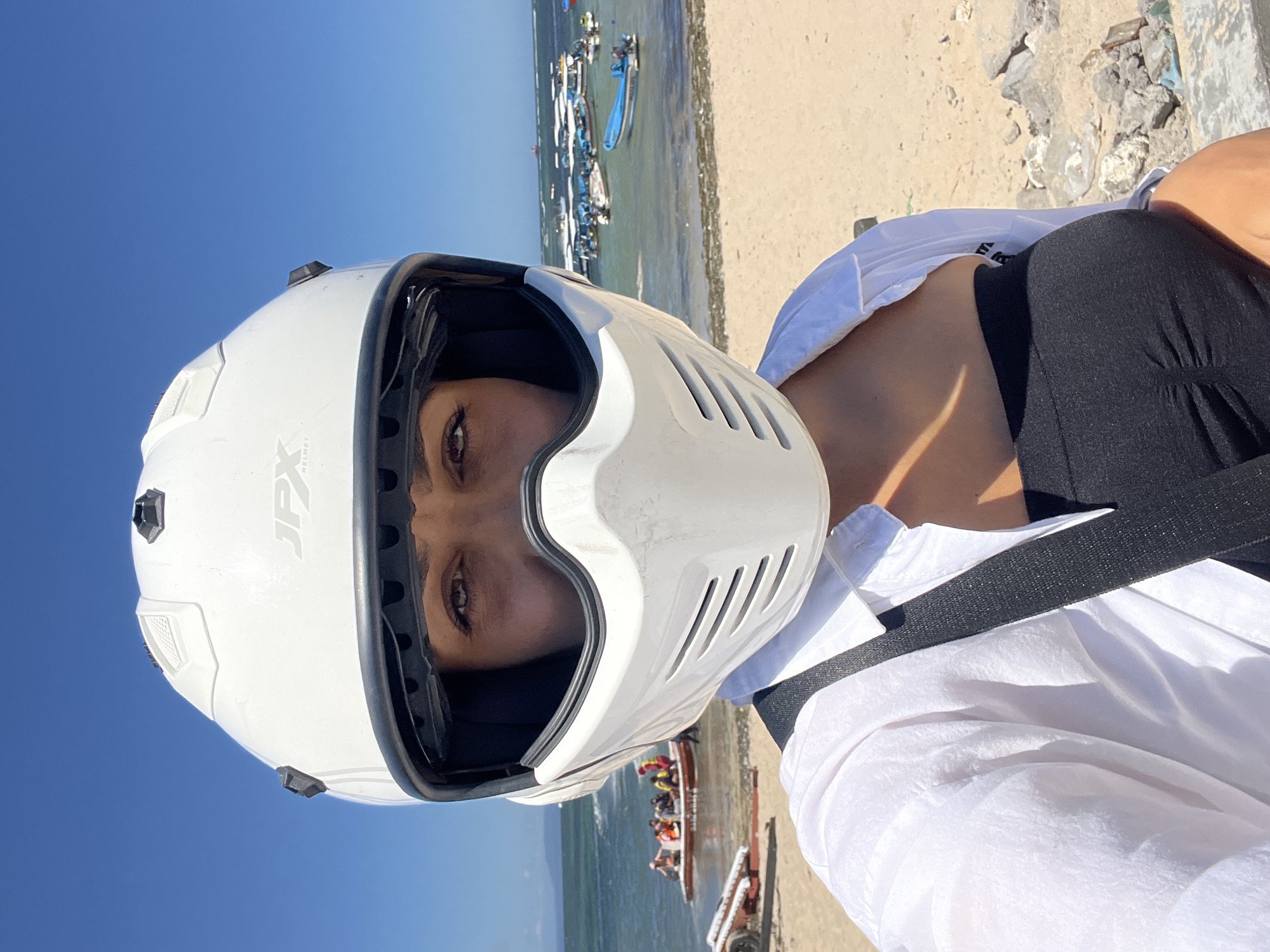 A woman wearing a white helmet stands on a beach, with her face visible through the helmet's opening. She is wearing a white shirt and black top underneath. Behind her is the ocean, a sandy beach, and several boats. The location is Unknown, Indonesia.