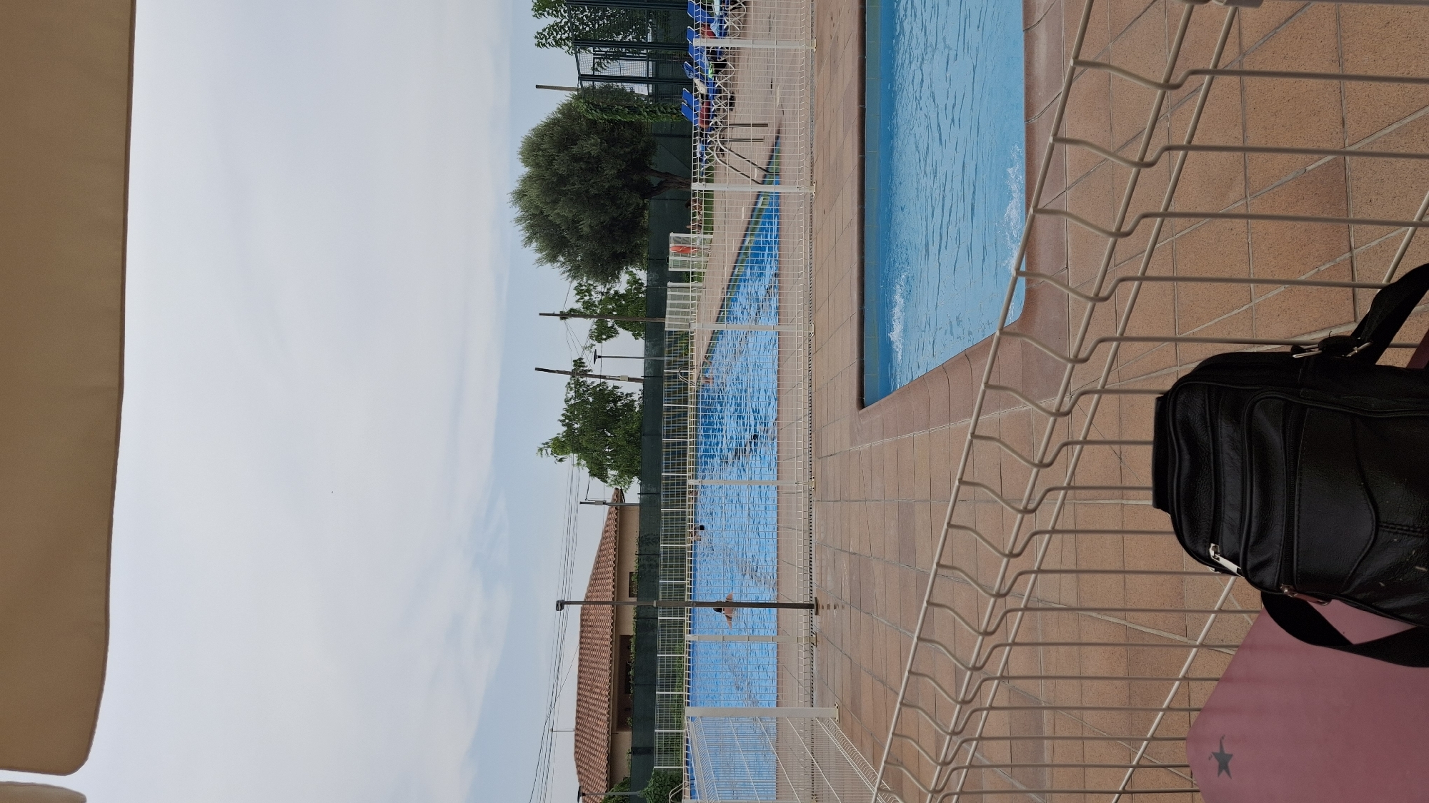 The image shows a partially fenced outdoor swimming pool area. In the foreground, a black leather shoulder bag rests on a pink surface, partially obscured by a white metal fence. Beyond the fence, a large blue swimming pool occupies the middle ground, with several people swimming. A tiled walkway surrounds the pool, with a second, smaller pool visible in the lower right corner. In the background, a red-tiled roof building and lush green trees are visible under a cloudy sky. The location is identified as the municipal swimming pool in La Fresneda, Spain.