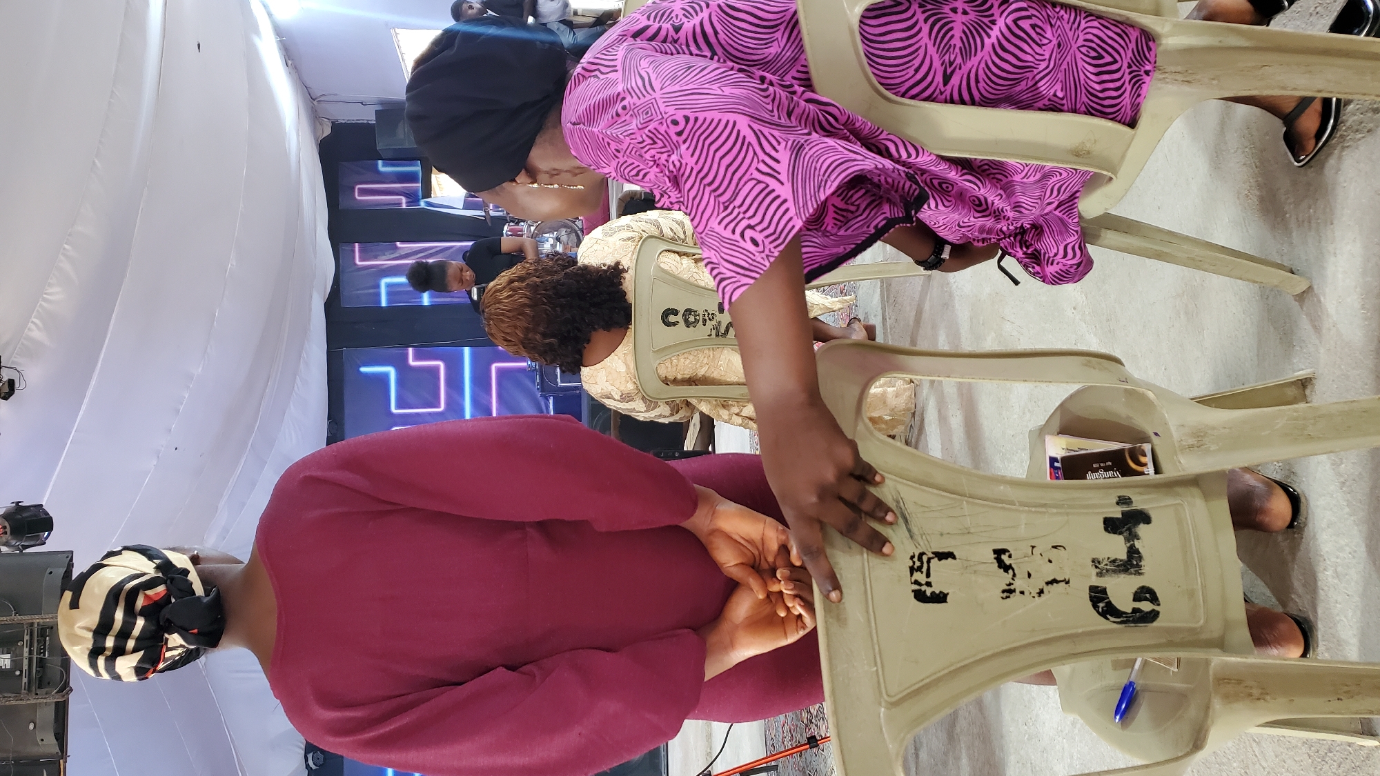 This image depicts an indoor gathering, likely a church service or event, in Jalingo, Nigeria. In the foreground, a woman from the back is wearing a maroon dress and a patterned headscarf. To her right, another woman in a pink and black patterned dress is seated, facing away from the camera, and resting her hand on a plastic chair. This chair, also in the foreground, is light beige with black markings on its back, and a blue pen and some pamphlets are visible on it.
Further in the background, more people are seated on similar chairs. One woman in a gold dress is visible from the back, and a person in a black outfit is standing near what appears to be a stage with blue and pink neon lights. The environment seems to be a large indoor space with a white draped ceiling. The overall atmosphere suggests a community gathering.