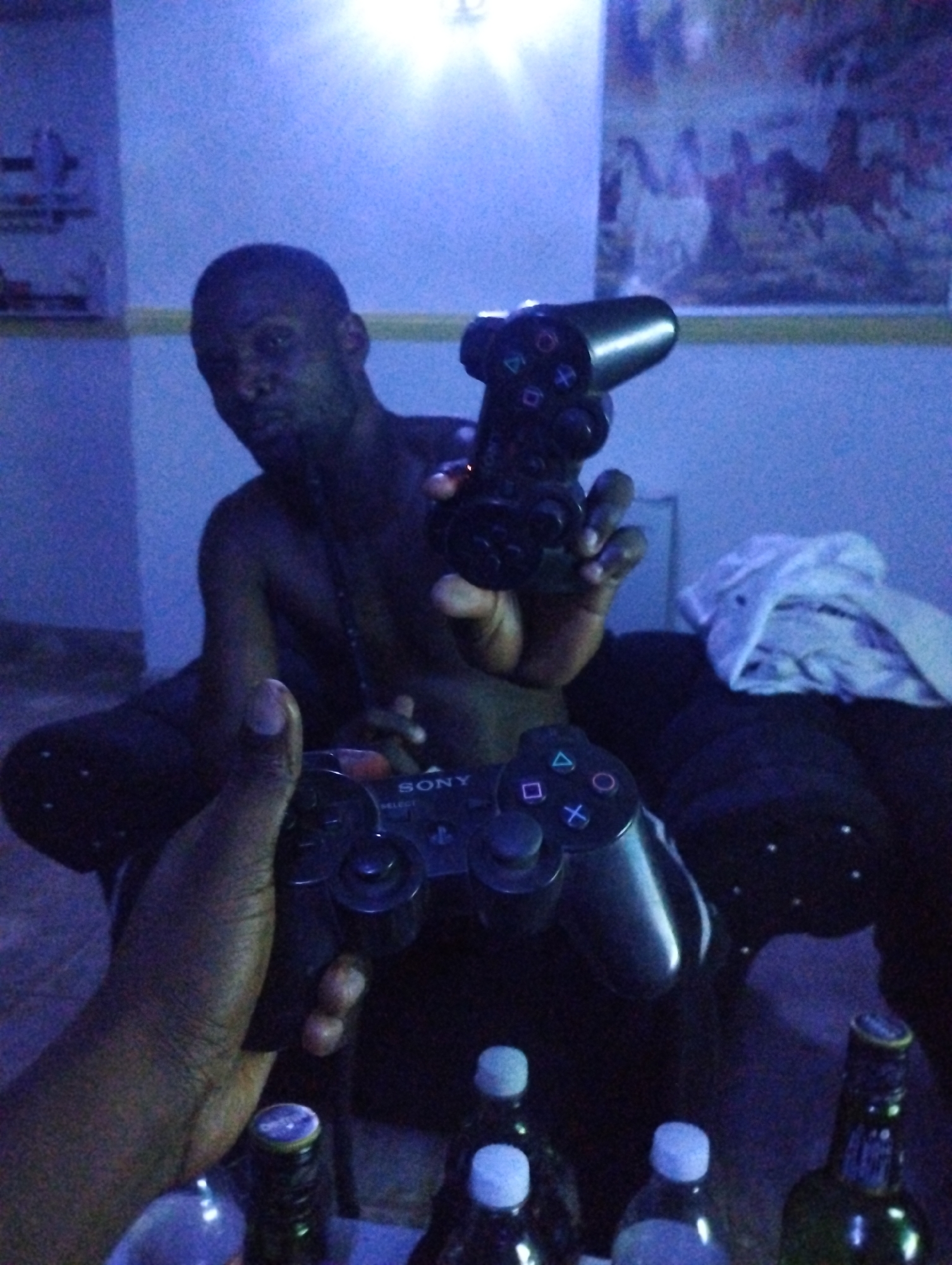 An African man is sitting in a dark room, holding two Playstation controllers. Four glass bottles with caps are on a table in the foreground. A painting is visible on the wall behind him. The location is Tumfure, Nigeria.