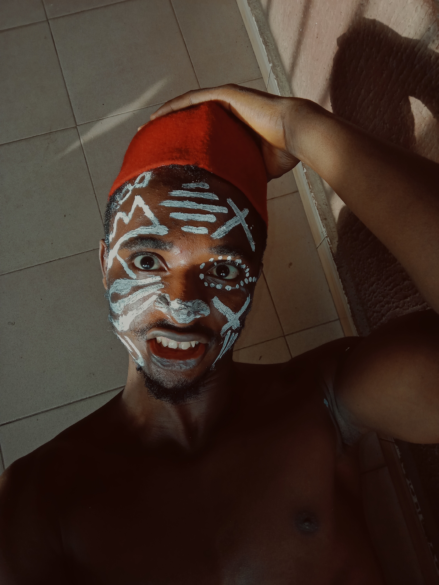 A shirtless, dark-skinned man is depicted from a low-angle, selfie perspective, wearing a red fez and adorned with white face paint. The paint consists of horizontal lines on his forehead, abstract shapes around his eyes, and lines framing his mouth and beard. He's holding the fez with his right hand, baring his teeth in a slightly theatrical manner. The background consists of light-colored tiled flooring and a wall, partially illuminated by sunlight. The scene is in a space that appears to be indoors. There is no specific evidence within the image to place it within Nkpor, Nigeria.