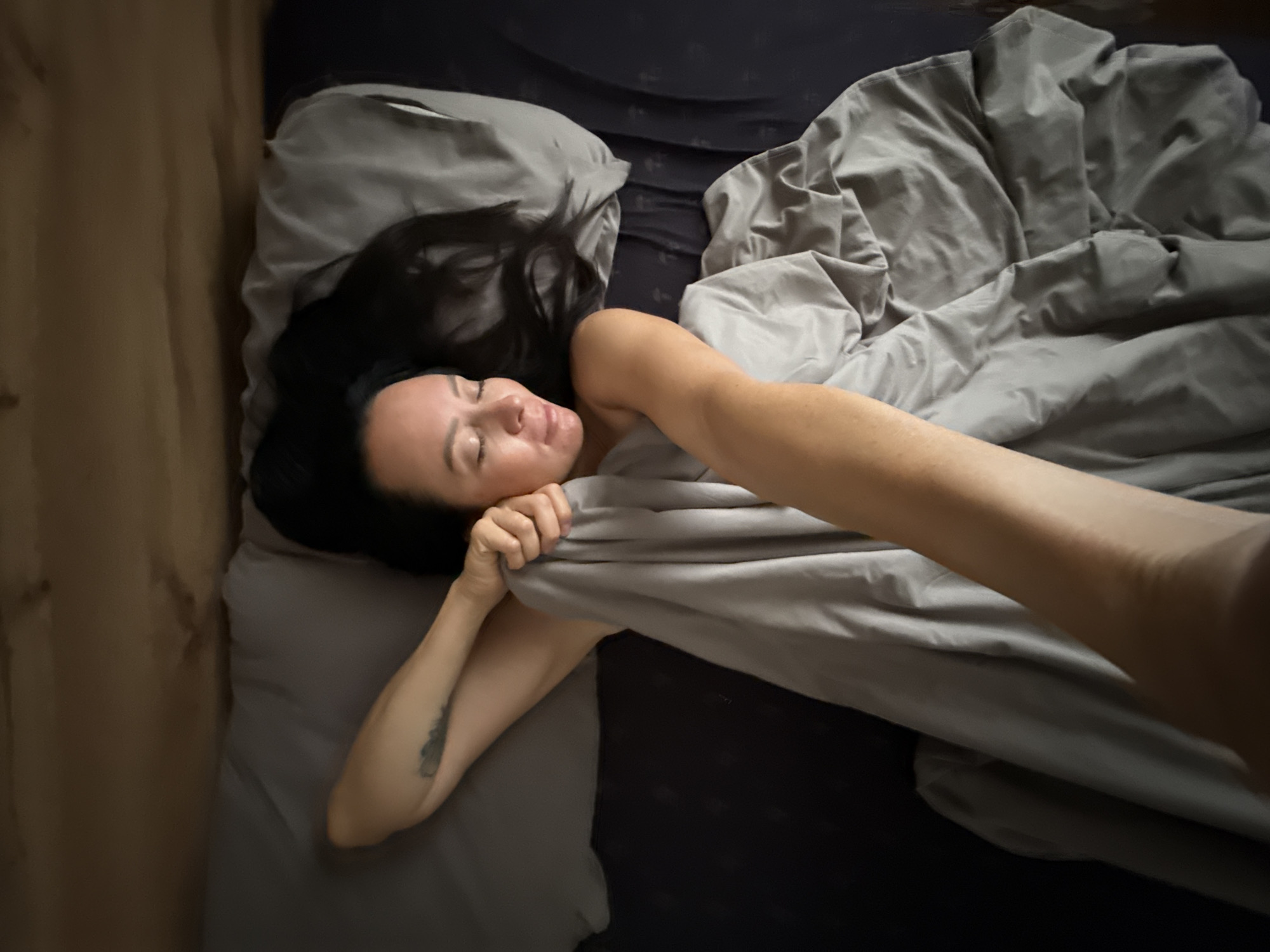 A woman with dark hair lies in bed, her eyes closed and her head resting on a grey pillow. Her left arm is raised, and her hand is near her face, pulling a grey blanket. The woman has a tattoo on her left upper arm. A person's arm, held towards the viewer, is visible in the foreground, partially covered by the grey blanket. The background consists of bedding and a wooden headboard. This appears to be a domestic bedroom scene. There is no information available to determine the location in the city of Chaikovsky, Russia.