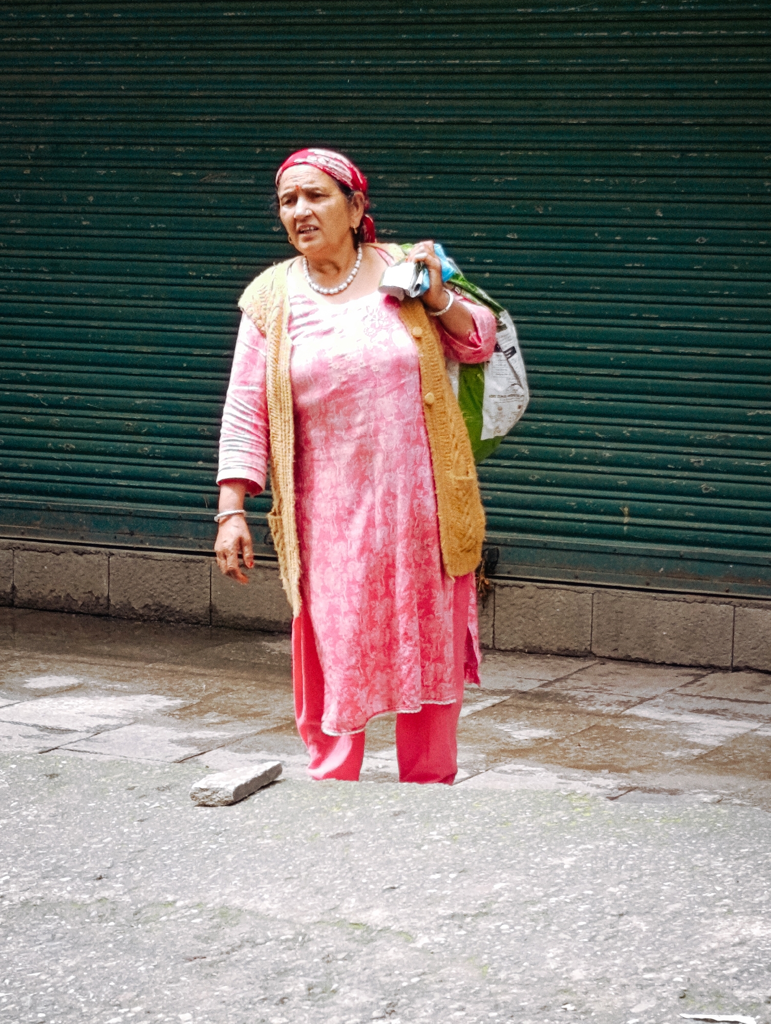 A woman in Vashisht, India, stands in front of a closed green metal shutter. She wears a pink patterned tunic with a mustard-colored knitted vest over it and a red headscarf. She carries a shopping bag over her left shoulder. The ground in front of her is wet and appears to be made of stone tiles. A small rock rests on the ground in the foreground.