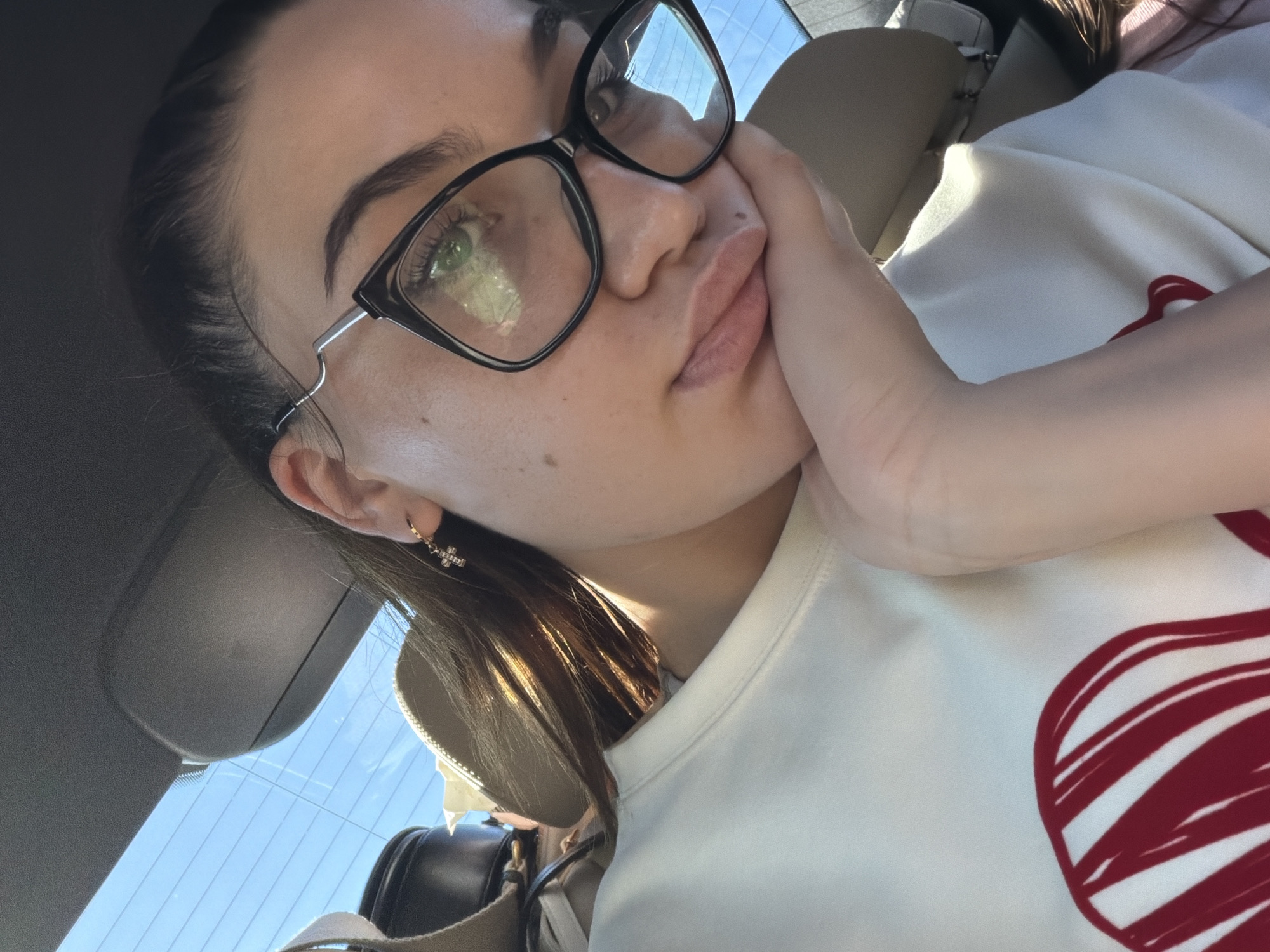 The image is a close-up, selfie-style shot of a young woman wearing glasses and a white sweatshirt with a red design on it. She is resting her chin on her hand, with her eyes looking down and to the side. The background appears to be the interior of a car, with a view of the sky through the rear window. The lighting suggests it is daytime. There are no specific landmarks or indicators that place the image in Zorleni, Romania.