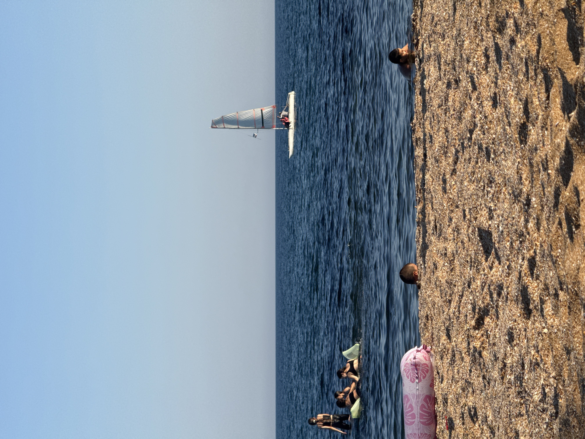A sunny day at the beach is captured in this vertical, eye-level shot. The foreground is dominated by a textured, shell-laden sandy beach. A long, pink inflatable with a floral pattern rests on the sand, partially submerged in the water at the left edge of the frame. To the right, a young boy's head and shoulders are visible as he partially submerges in the water. The middle ground features the calm, deep blue sea. Several people are present in the water. On the left, three individuals are in the water, two of them on yellow inflatable mats. A single person stands in the water to their left. Further out in the water, a white catamaran with red accents sails across the frame, carrying several passengers. The sky above is a clear, pale blue, indicating fair weather.