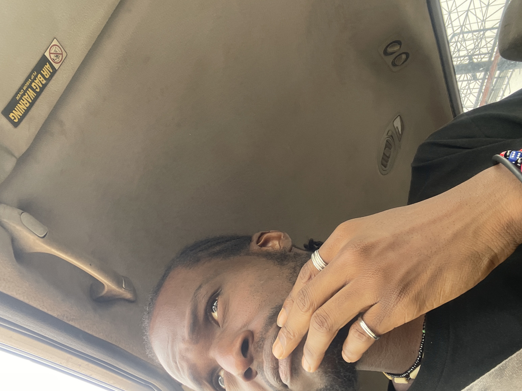 The image shows a close-up, low-angle shot of a man's face and hand inside a vehicle. The man is looking upwards and to the right, with his right hand resting against his mouth. He is wearing a black t-shirt and a necklace with black and white beads. Two silver rings are visible on his fingers. The interior of the vehicle is visible, including the ceiling with air vents and a grab handle. A warning sticker for the airbag system is present on the ceiling. The background suggests an urban environment with industrial structures visible through the window. The lighting is natural, possibly from the window. The image does not contain any specific details that would allow for the identification of the location as Jos, Nigeria.