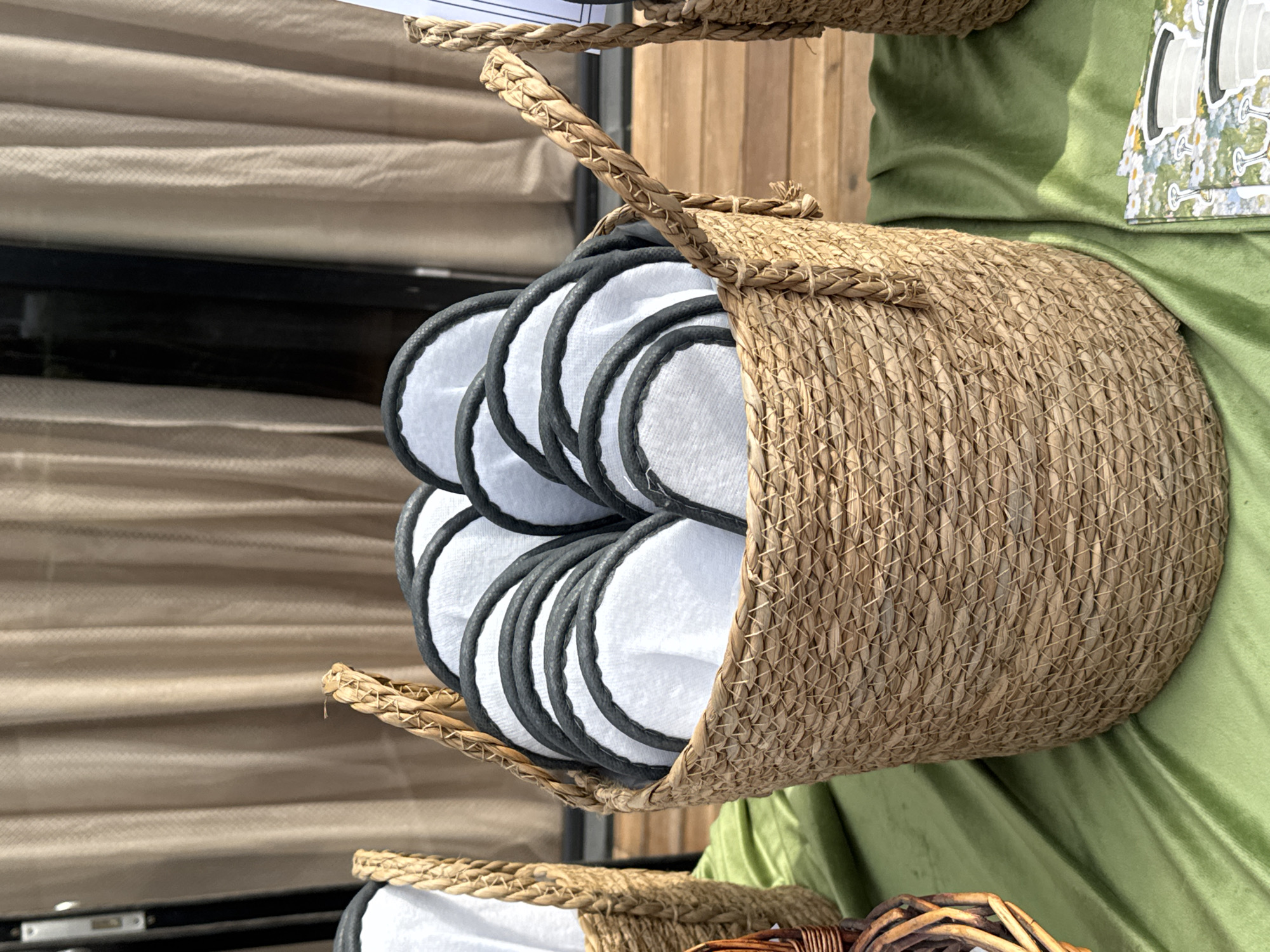 A close-up, eye-level shot shows a woven straw basket filled with white round pads featuring black scalloped edges, likely reusable makeup remover pads. The basket is placed on a green satin cloth laid out on a wooden deck. In the background, a light brown curtain is visible, partially obscuring a dark, reflective surface that could be a window or door. The scene appears to be outdoors, with natural light casting subtle shadows. The location is Arashan, Kyrgyzstan, suggesting a rustic or natural setting.