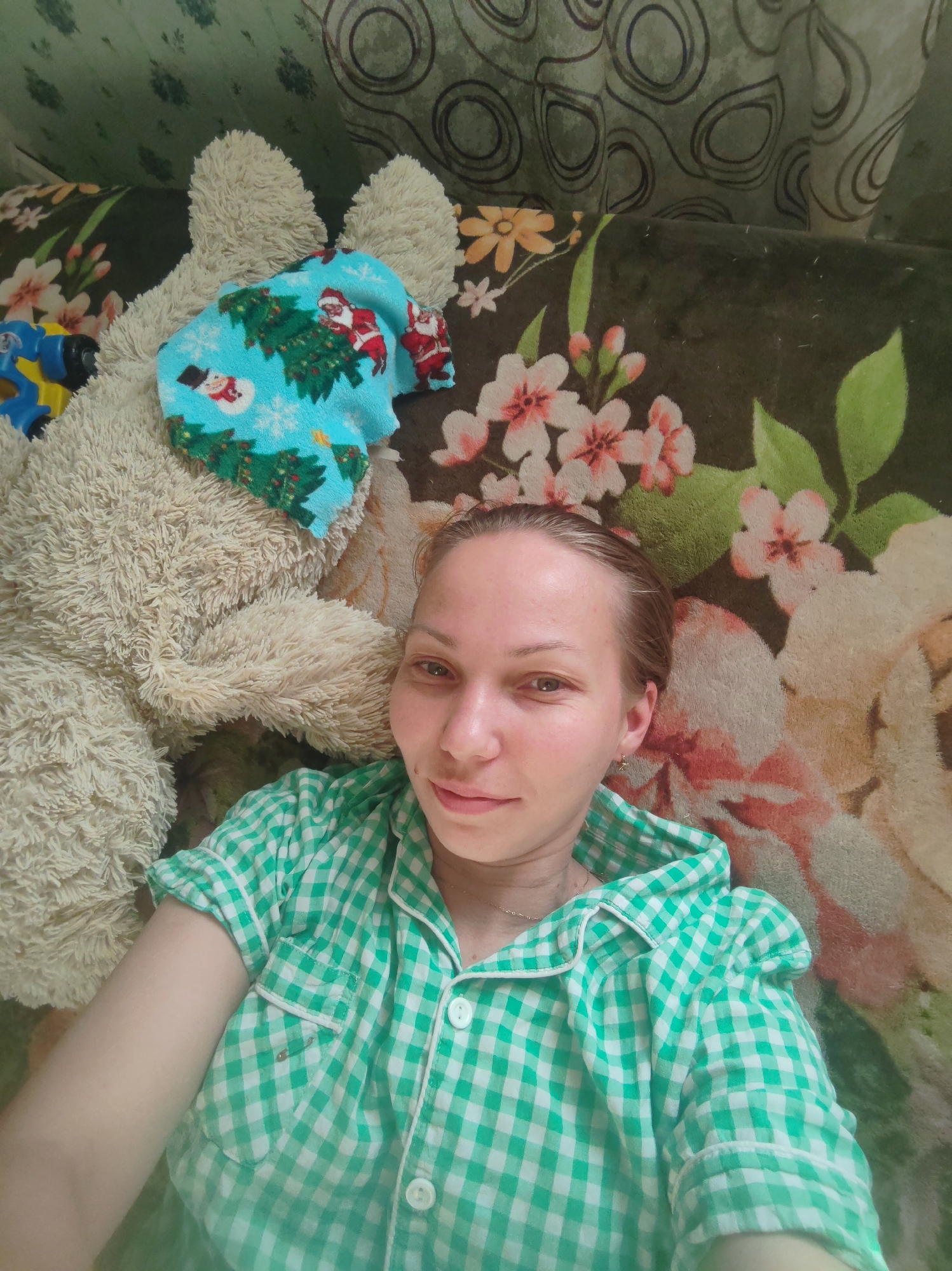A woman is lying down, close to a large, tan stuffed bear. The woman is wearing a green and white checkered pajama top with a collar and buttons. A blue cloth with images of Santa Claus and Christmas trees is draped on the bear. A blue and yellow toy is also near the bear. The background includes a floral patterned blanket and a patterned curtain. The location is unknown and cannot be determined to be in Чухлэм, Russia.