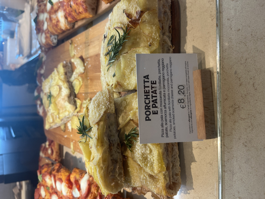 This image captures a close-up of a food display counter, likely in a bakery, pizzeria, or deli in Dorno, Italy. The central focus is a rectangular pizza, cut into several slices, prominently featured on a wooden cutting board. This pizza, labeled "PORCHETTA E PATATE," is topped with thin, lightly browned potato slices, visible chunks of porchetta, fresh sprigs of rosemary, and a sprinkling of grated cheese.
To the bottom left and in the background, other rectangular pizza slices are partially visible, one featuring a red sauce base with melted white cheese, suggesting a classic margherita or similar variety. The food is displayed under bright, likely artificial, lighting.
A white informational card stands next to the potato and porchetta pizza. The text on the card describes the dish in both Italian and English: "PORCHETTA E PATATE," "Pizza alla pala con mozzarella fiordilatte, porchetta, patate, scamorza affumicata e parmigiano reggiano" and "Pizza alla pala with fiordilatte mozzarella, porchetta, potatoes, smoked scamorza cheese and parmigiano reggiano." The price displayed is "€ 8,20." Additional text indicates that ingredients and allergens are listed on ingredient signs, and that the product's base was frozen at source.
No people are visible in the frame. The setting appears clean and organized, with the pizzas presented appealingly for sale. The time of day is not explicitly indicated but the bright lighting suggests daytime business hours.