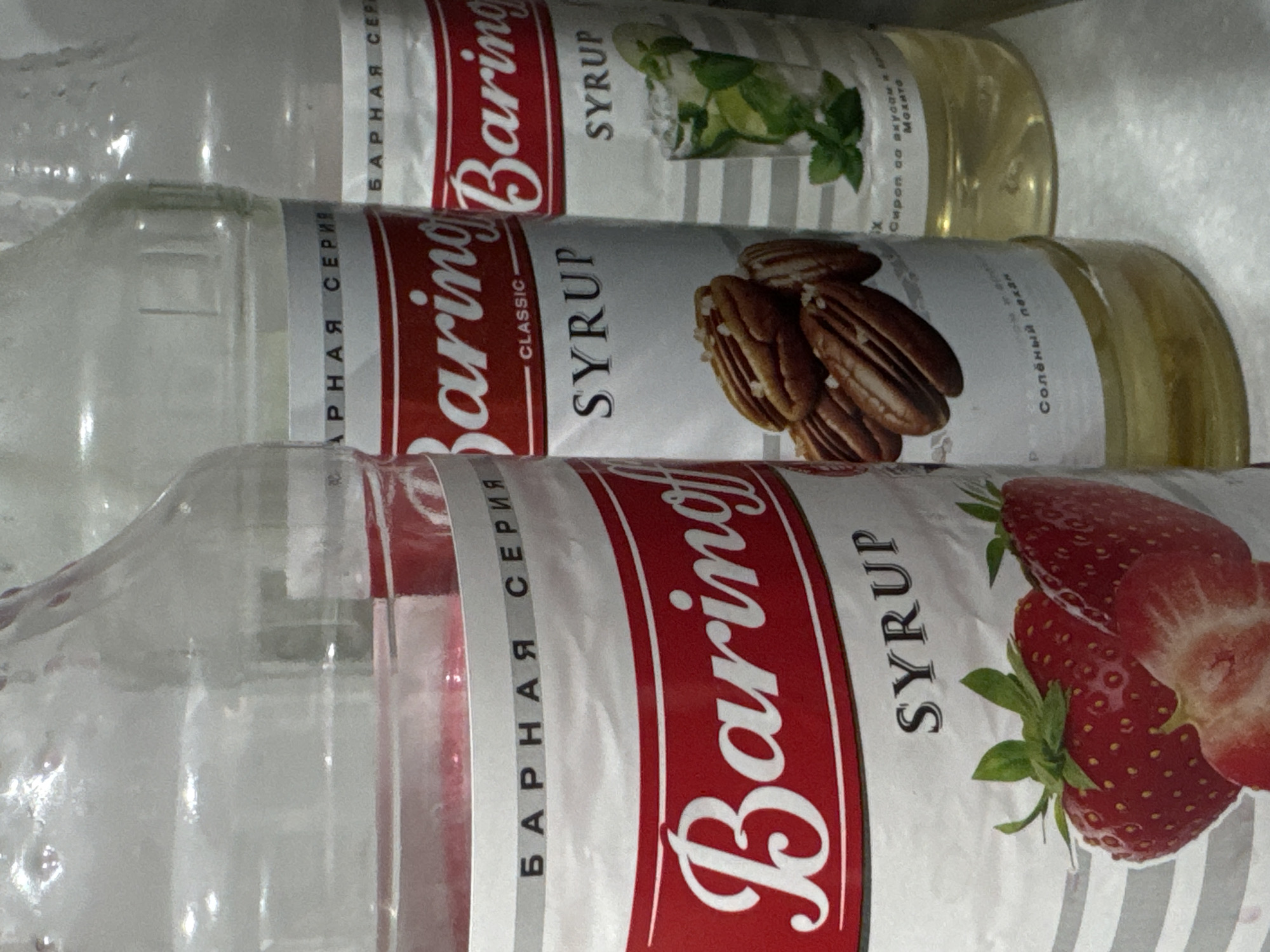 The image shows three bottles of syrup. The bottles are clear plastic and contain a light yellow liquid. The labels on the bottles are white with red and gray accents. The labels feature the brand name "Baringoff" in large white cursive letters. The label on the leftmost bottle also depicts an illustration of strawberries. The middle bottle's label has an illustration of pecans and the text "CLASSIC SYRUP" and "Солёный пекан" (Salted Pecan). The rightmost bottle's label shows an illustration of a drink with mint and lime and the text "SYRUP" and "Сироп со вкусом Мохито" (Mojito flavored syrup). The bottles are arranged in a row on a light-colored surface, possibly a shelf or countertop. The background is slightly blurred but appears to be an indoor setting. The image is taken from a slightly low angle, looking up at the bottles. There are no people visible in the image. This scene is likely in a commercial establishment or a home kitchen in Mikhaylovsk, Russia, given the Russian text on the labels.