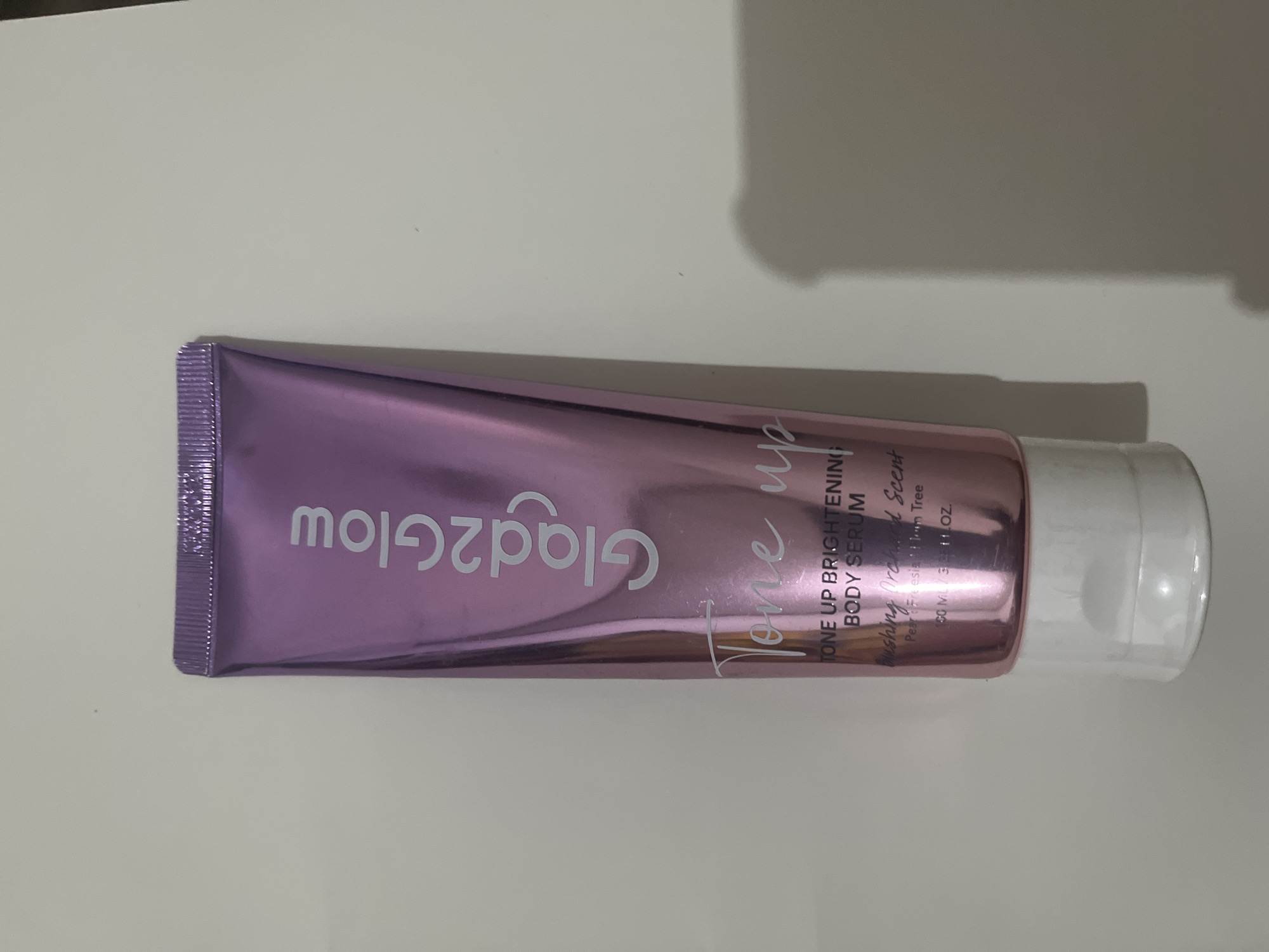 A metallic pink tube of "Tone Up Brightening Body Serum" by Glad2Glow is shown against a plain, light-colored background. The tube is upright and centered in the frame, with its cap at the bottom. Text on the tube reads "Glad2Glow", "Tone up", "TONE UP BRIGHTENING BODY SERUM", "Blushing Orchard Scent", "Pear, Freesia, Plum Tree", and "50 ML / 1.7 fl. OZ.". The background is featureless and appears to be a wall or tabletop. There is a shadow cast to the right of the tube. The image does not contain any information about the city of Meliau Hulu, Indonesia.