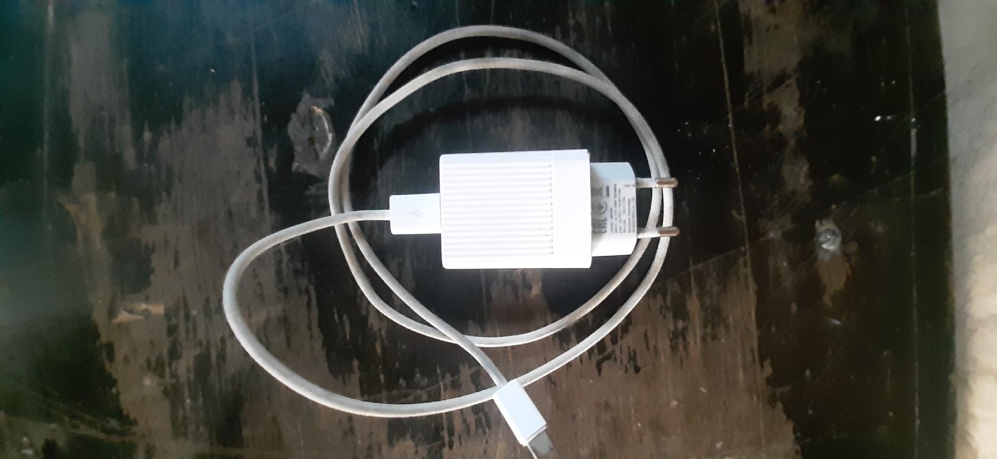 The image shows a white USB charger with a white USB cable. The charger is plugged into the cable, and the cable is coiled loosely around the charger. The charger is made of white plastic and has a ribbed texture. The cable is also white and appears to be made of rubber or silicone. The charger has two metal prongs that would plug into a wall outlet, and a USB port. There is text on the charger, but it is too small to read. The charger and cable are lying on a dark, wooden surface that appears to be a table. The surface is scratched and worn in places. The lighting in the image is dim, and there is no indication of the time of day or weather. The overall scene is simple and uncluttered, with the focus solely on the charger and cable. There are no people or other objects visible in the image. The setting appears to be indoors.