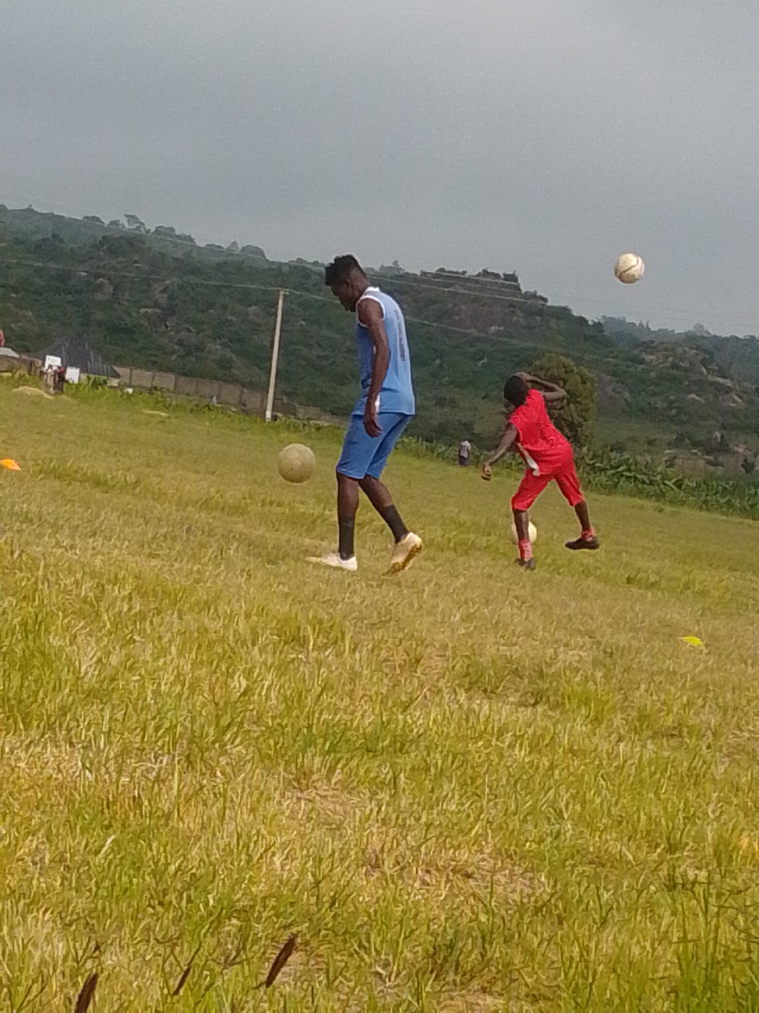 The image shows two young males playing soccer on a grassy hill under an overcast sky. The person on the left is wearing a blue jersey and blue shorts, and is kicking a soccer ball. The person on the right is wearing a red jersey and red shorts, and appears to be running. In the background, there are trees and hills, suggesting a rural or undeveloped area. The ground is covered in dry grass. There are some orange and yellow markers on the grass, likely for a training exercise. The overall scene depicts a casual outdoor activity.