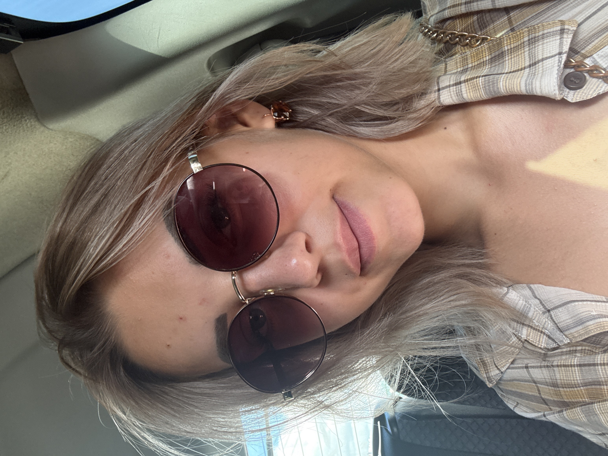 A young woman with blonde hair, wearing sunglasses and a plaid shirt, is photographed from the chest up. She is seated in what appears to be the passenger seat of a vehicle. The background shows the interior of a car, with a window on the left side of the frame, partially obscured by the woman's hair. The lighting suggests it is daytime, with sunlight casting shadows on her face and shoulder. The image does not contain any specific landmarks or features that would allow for the determination of its location in Yuzhno-Sakhalinsk, Russia.