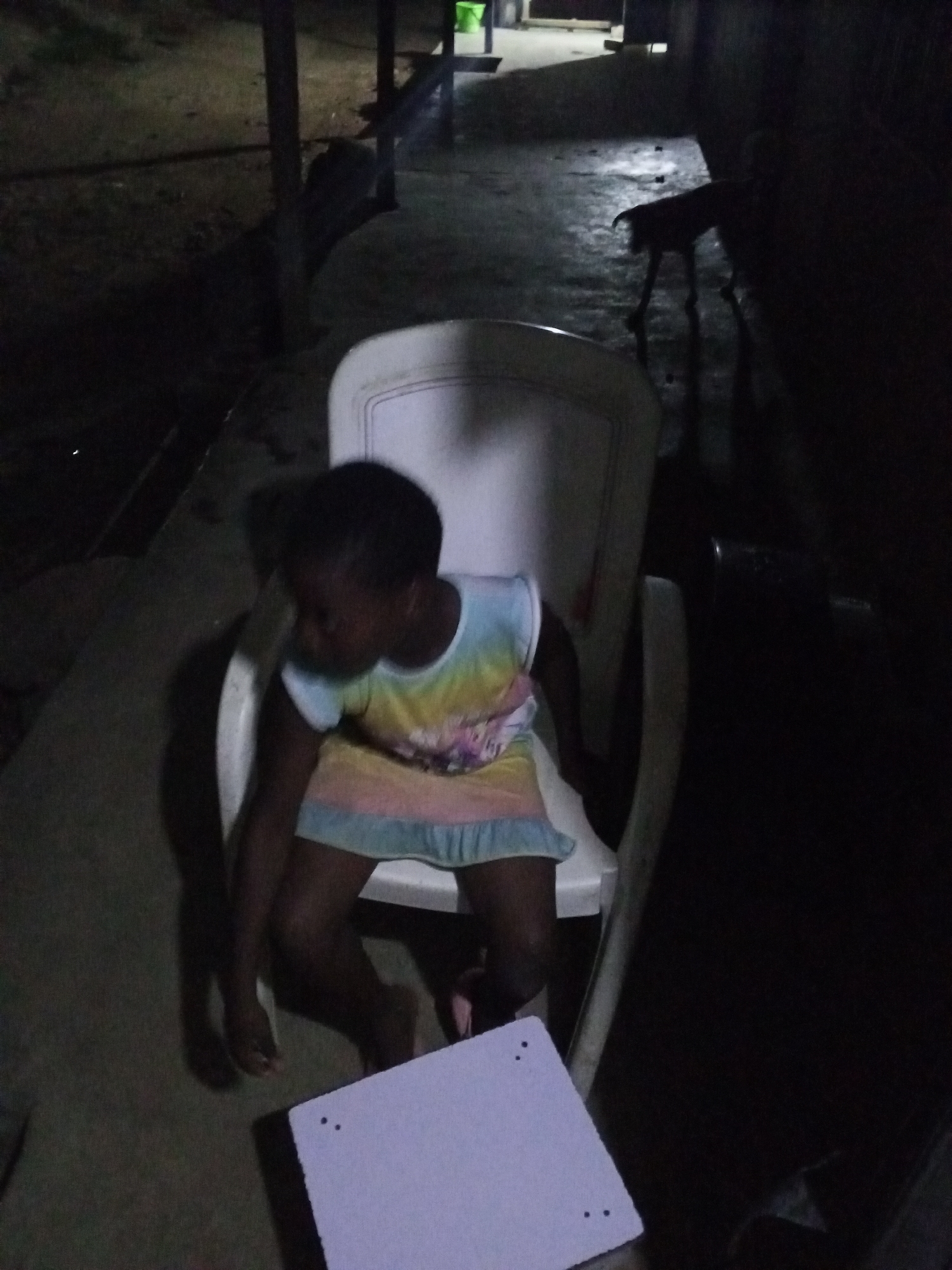 A young child is sitting on a white plastic chair outside in the dark. The child wears a light-colored dress with a rainbow design. A small, square white object with dark spots on its surface rests on the ground near the child's feet. In the background, indistinct shapes suggest a building structure and foliage. A dark, animal-like form can be seen standing in the distance. The location is identified as Jos, Nigeria.