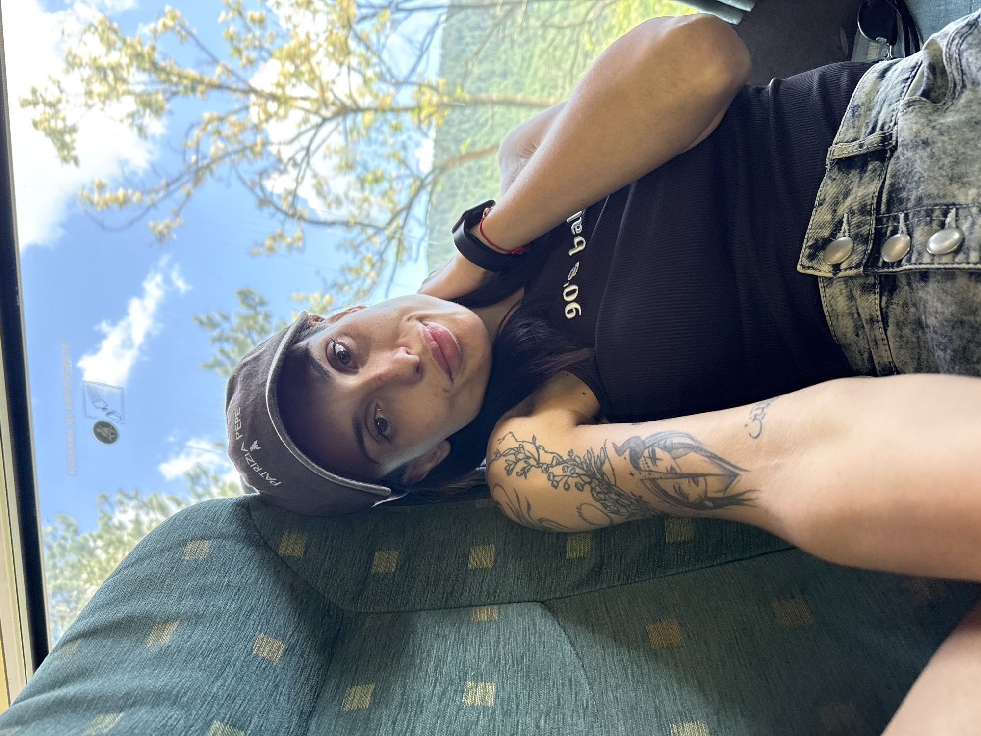 A person is lying down on what appears to be a bus seat. The seat has a green patterned upholstery. The person is wearing a black tank top with the words "90's baby" on it, a gray cap with the name "Patrizia Pepe" on it, and a blue denim jacket with silver buttons. They have a black watch on their wrist and tattoos on their left arm. Through the window, the sky, trees and mountains can be seen. The image was taken in the city of Balgarene, Bulgaria.