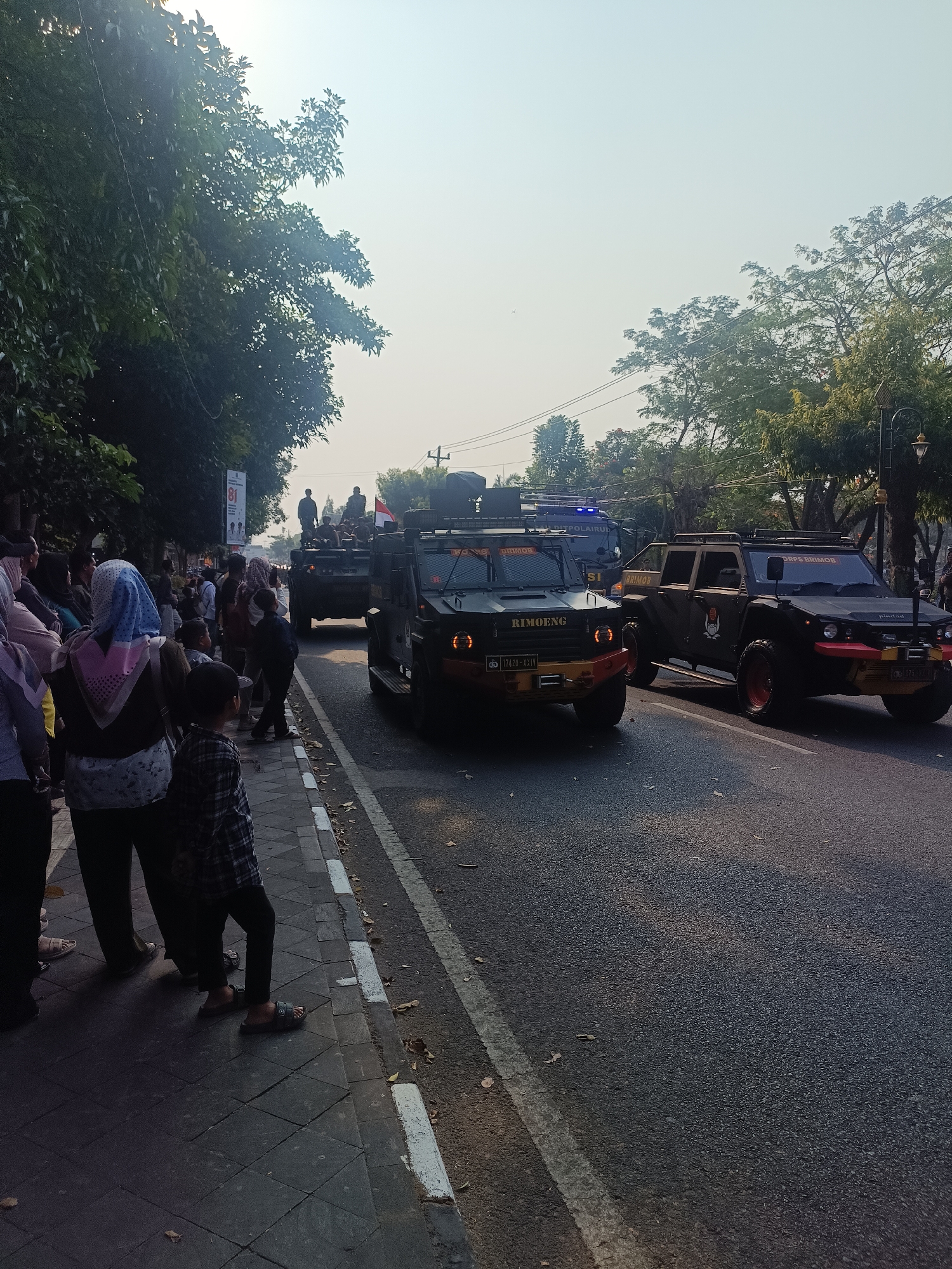 On a paved road in Sleman, Indonesia, an apparent convoy of armored vehicles is moving, with onlookers gathered on the sidewalk. The sky is bright, suggesting daytime. The foreground features a paved sidewalk with fallen leaves. A group of people, including women wearing headscarves and children, are standing and observing the vehicles. One child in a checkered shirt is positioned closest to the camera. The road itself is asphalt, with white lane markings.
The convoy consists of at least three military or police vehicles. The most prominent is a dark, rugged vehicle with large tires, a reinforced body, and what appear to be tactical lights. The words "BRIMOB" and "RIMBONG" are visible on its front. Another similar vehicle is behind it, and a third vehicle, a dark pickup truck-like vehicle with a crew cab, is to the right, also appearing to be part of the convoy. A flag, possibly Indonesian, is seen on one of the vehicles. In the background, trees line the street, and a bright, hazy sky is visible. A poster or banner with the number "81" is on a pole to the left, adjacent to a tree. The overall impression is of a public event or procession.