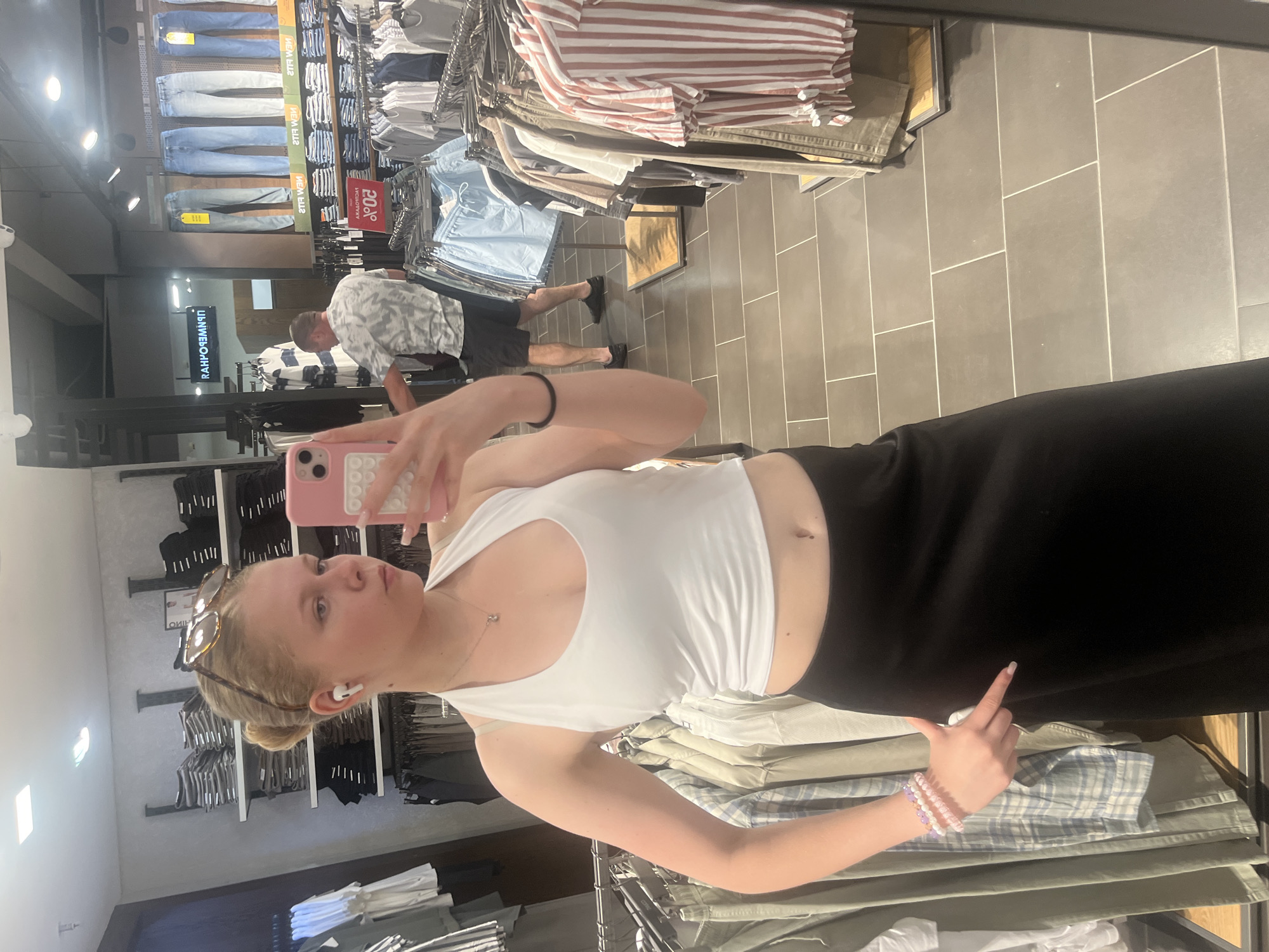 The image shows a young woman taking a mirror selfie in a clothing store. She is wearing a white crop top and a black skirt. She has blonde hair pulled back in a bun and is wearing sunglasses on top of her head and white earbuds. She is holding a pink phone in her right hand and is pointing her left index finger towards the camera with a playful gesture.
The store appears to be a men's clothing store, with racks of jeans, shirts, and trousers visible in the background. The floor is tiled with grey tiles. There are clothes on display, including a rack of jeans with "20%" sale tags. A sign with Cyrillic text is visible in the mirror reflection, indicating "RAHFOROЯM". The overall lighting suggests it is daytime. The woman's expression is relaxed and confident, suggesting a casual shopping experience. The location context provided is Minsk, Belarus.
