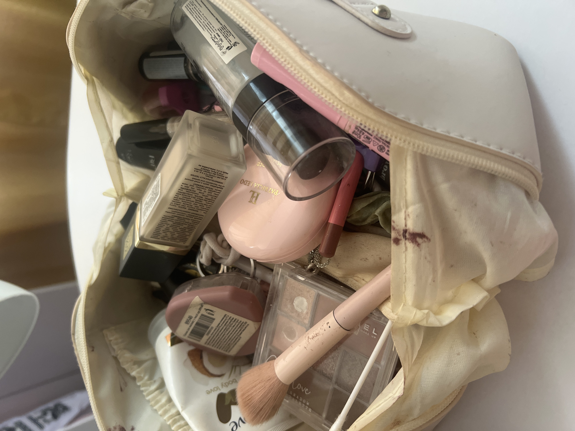 The image shows an open, cream-colored makeup bag filled with various cosmetics. The bag is on a white surface, possibly a table or counter. Inside the bag, visible items include a bottle of foundation, a pink makeup brush, an eyeshadow palette, a pink lip pencil, a tube of lotion or body cream, and several other unidentifiable cosmetic containers and accessories. Some of the items are partially obscured by others. The interior of the bag appears to have a floral pattern. There are also some dark stains on the inner lining of the bag. No people or animals are present in the image. The scene is indoors, and the lighting suggests it is daytime.