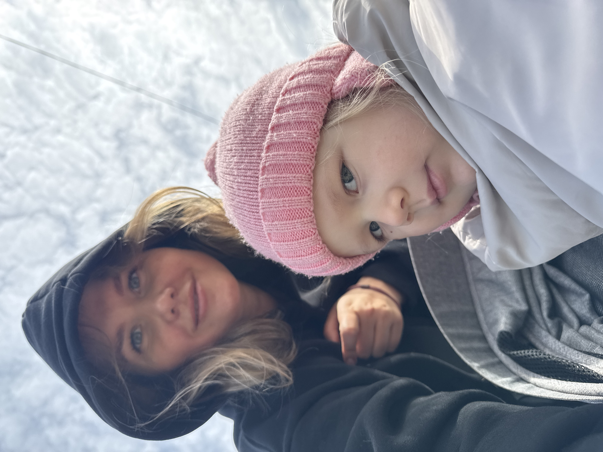 A selfie taken from a low angle features a woman and a young child. The woman, on the left, wears a black hooded sweatshirt, her blonde hair partially visible. The child, on the right, is wearing a pink knit hat and a gray and white jacket. The background shows a cloudy sky, possibly indicating an outdoor setting. The location could be Горный, Russia.