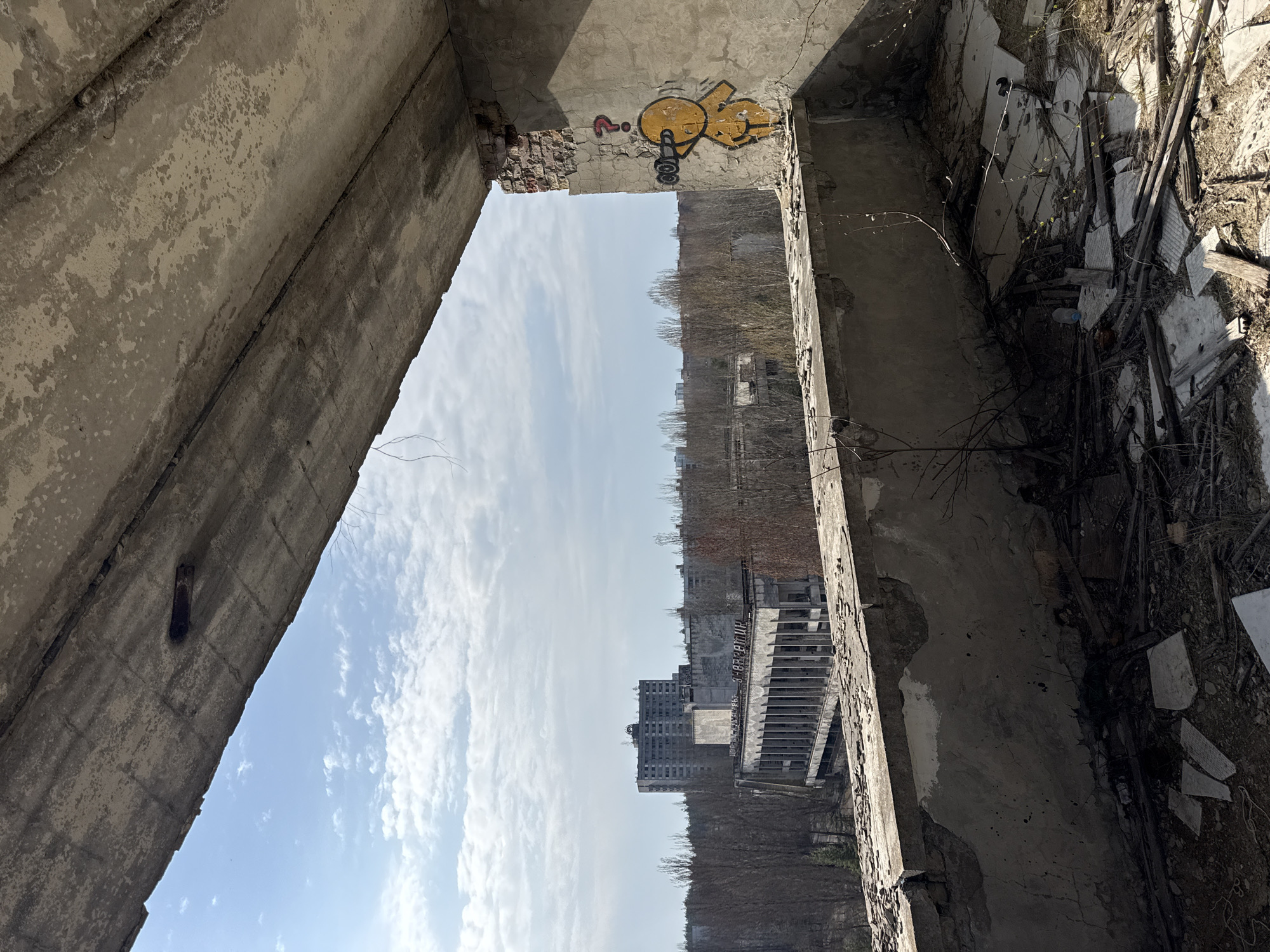 The image shows a view from within a dilapidated building in Prypiat, Ukraine. The decaying concrete structure frames the exterior view, showcasing other ruined buildings against a pale blue sky with scattered clouds. Inside, remnants of debris and shattered building materials litter the ground. Graffiti adorns the decaying walls of the building, adding to the sense of abandonment and decay.