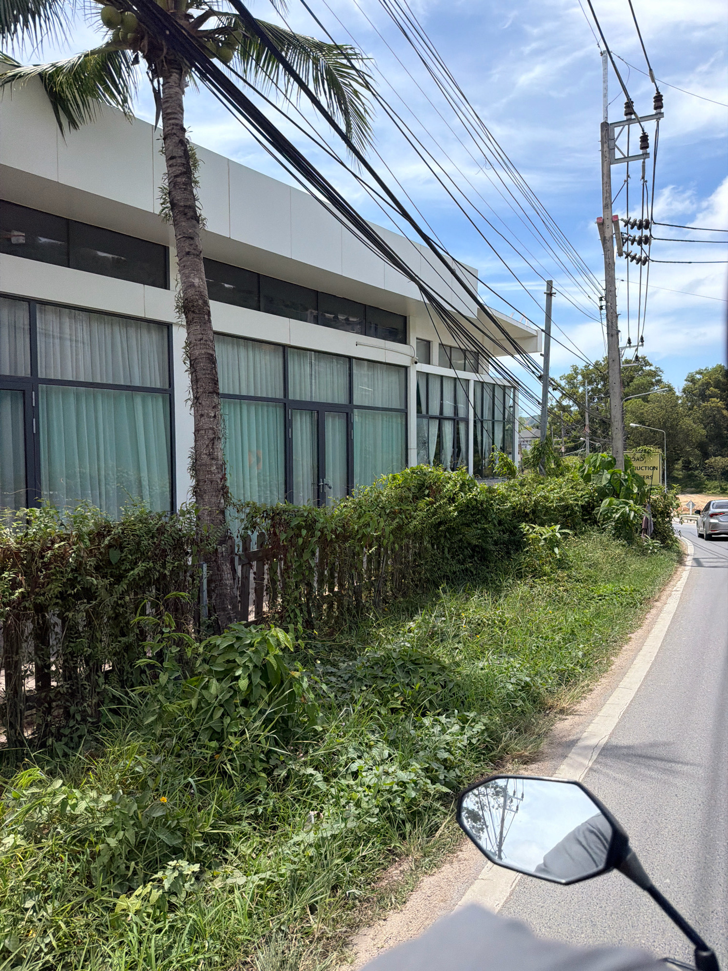 The image depicts a roadside scene in Choeng Thale, Thailand, on a bright, partly cloudy day. A modern, white building with large, curtained windows runs along the left side of the frame. In front of the building is a palm tree with coconuts and a wooden fence overgrown with dense green foliage. The road is on the right, with a white line marking its edge. A motorcycle's rearview mirror is visible in the lower right corner, reflecting the sky and utility poles. Across the road, there are more trees and a yellow sign. A silver car is visible further down the road, moving away from the viewer. The environment appears to be a semi-urban or suburban area with significant vegetation.